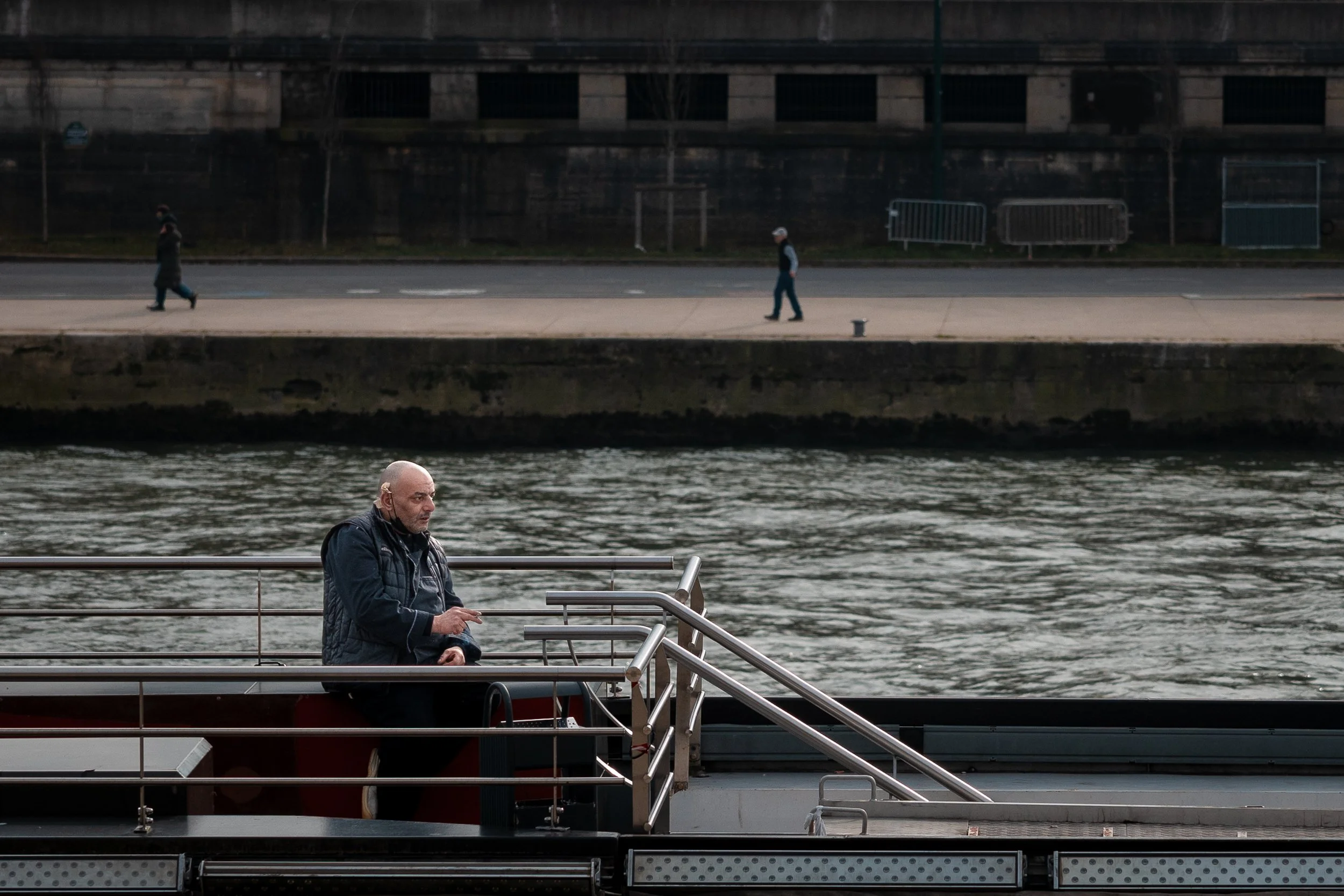A man with a bald head and grey beard sitting on a boat dock near the river, looking at his phone, with two people walking along the riverbank in the background.