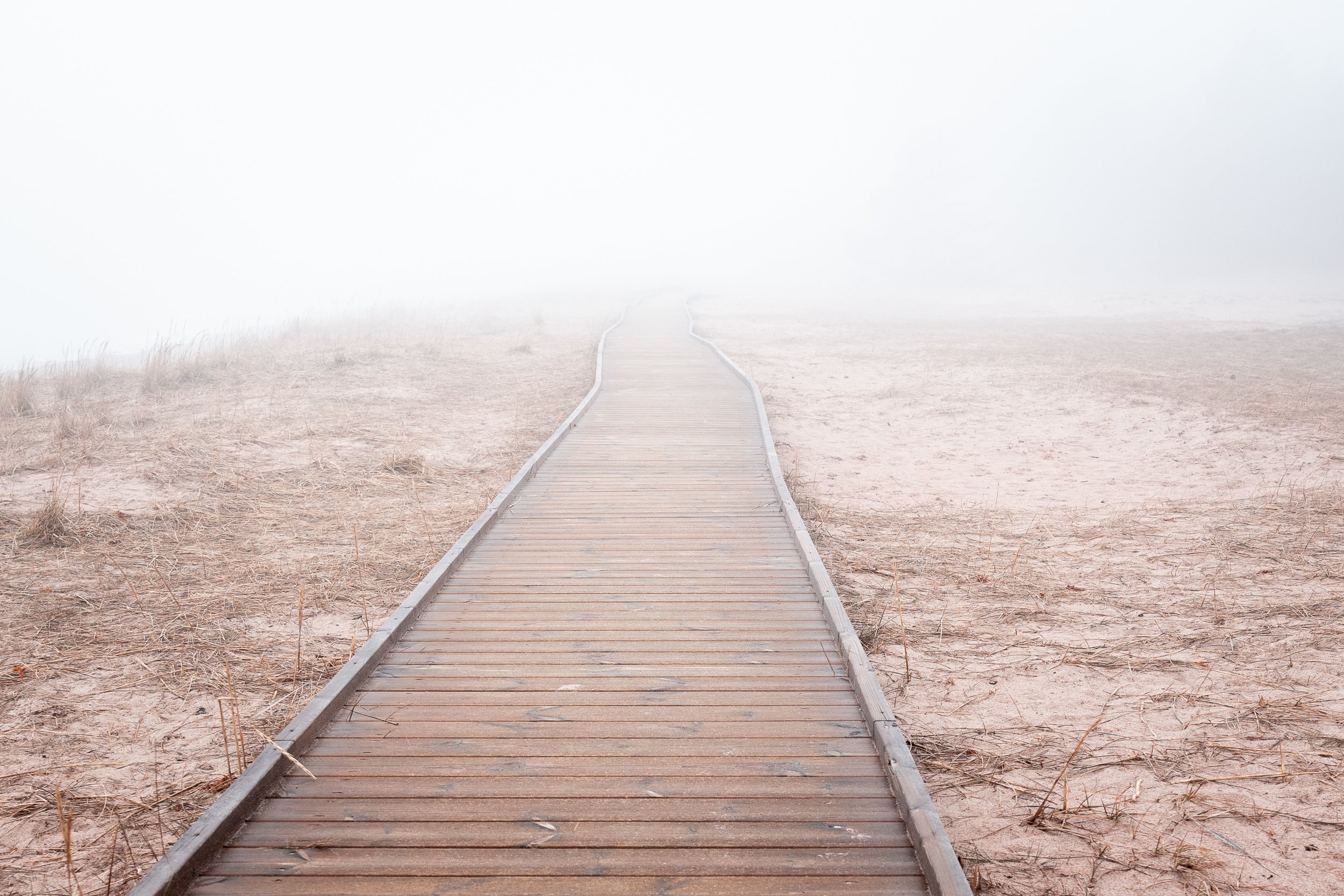 A wooden boardwalk fading into the foggy distance on a sandy beach with dry grass on either side.