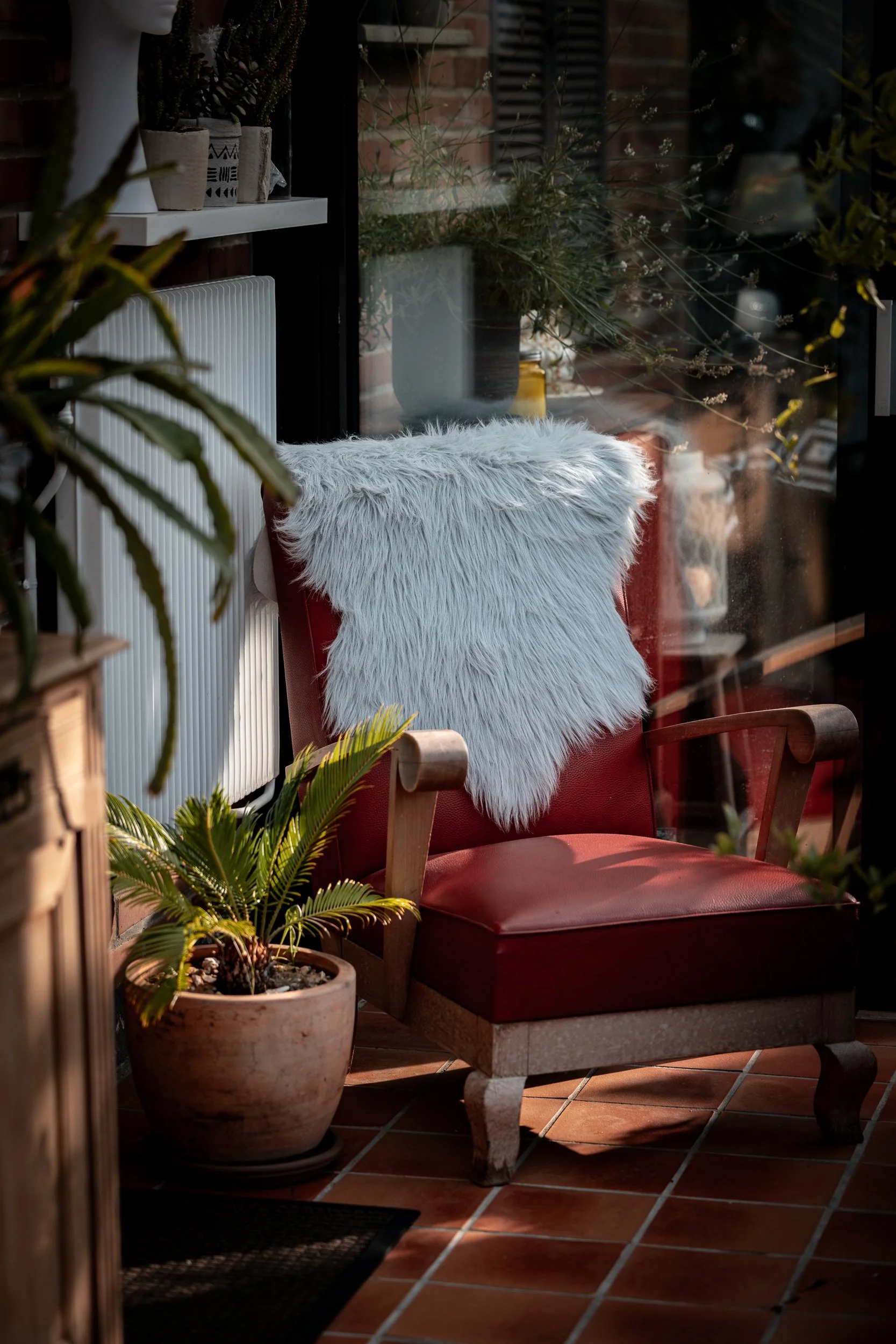 A cozy red armchair with a white faux fur throw draped over the back, situated indoors near a large glass window with plants and decorative items on a shelf, sunlight casting warm light on the terracotta tiled floor.