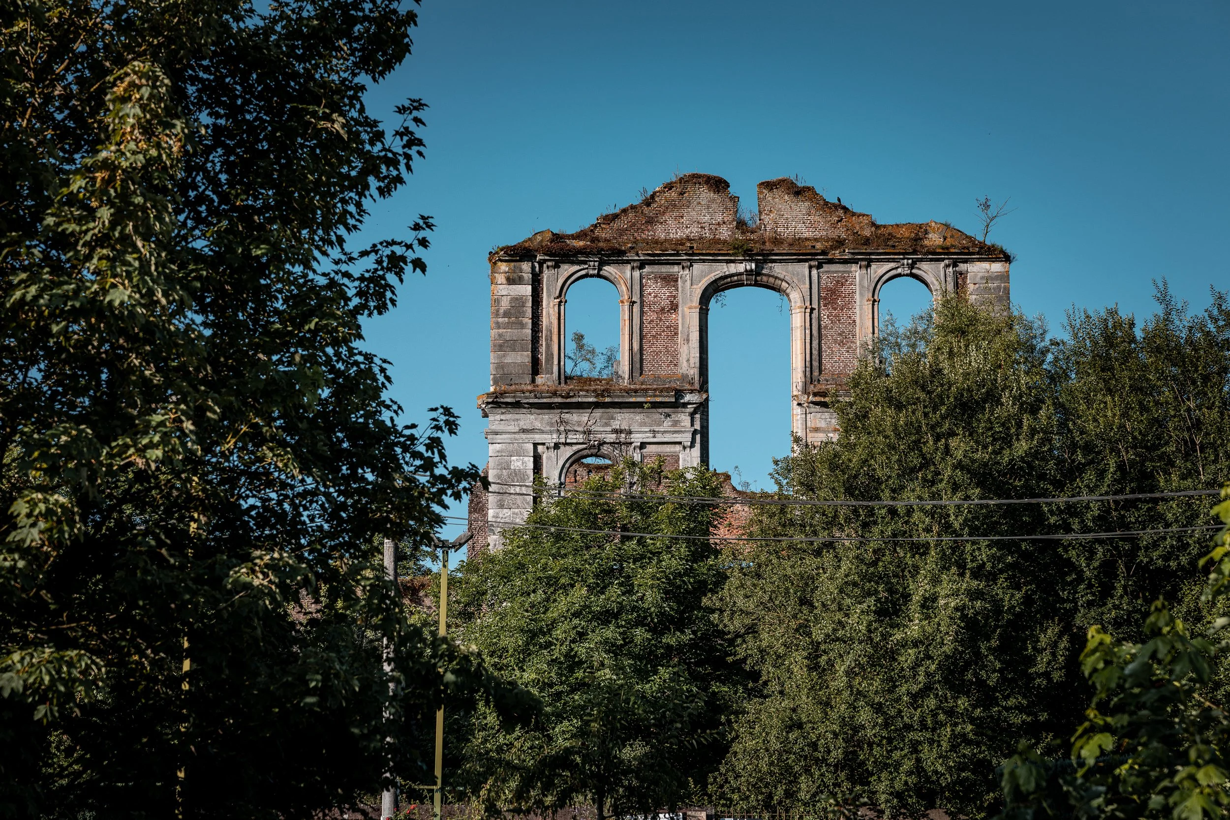 Ruins of an old brick and stone building with three large arches, partially destroyed, surrounded by green trees against a clear blue sky.