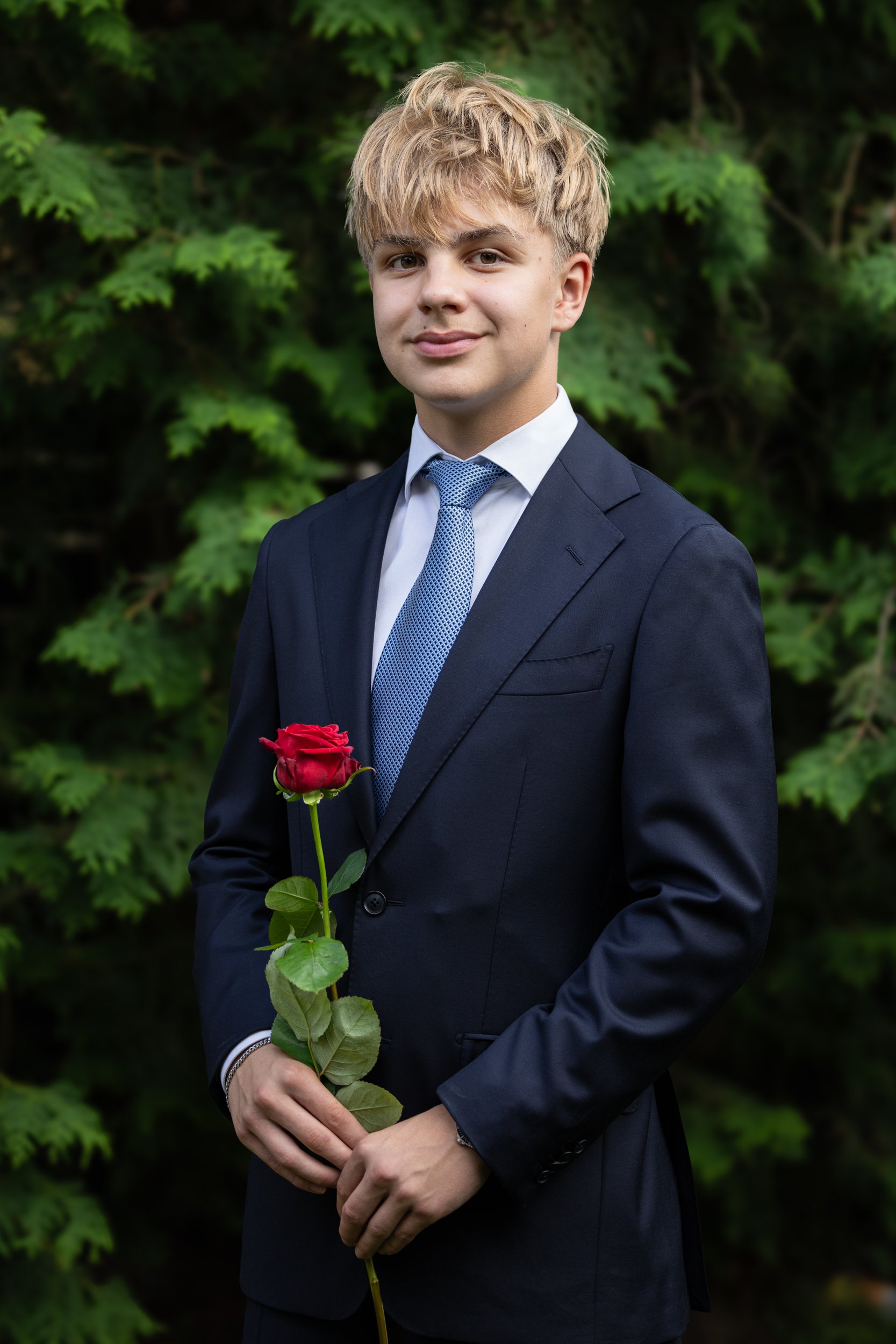A young man in a dark suit holding a red rose, standing outdoors with green foliage background.