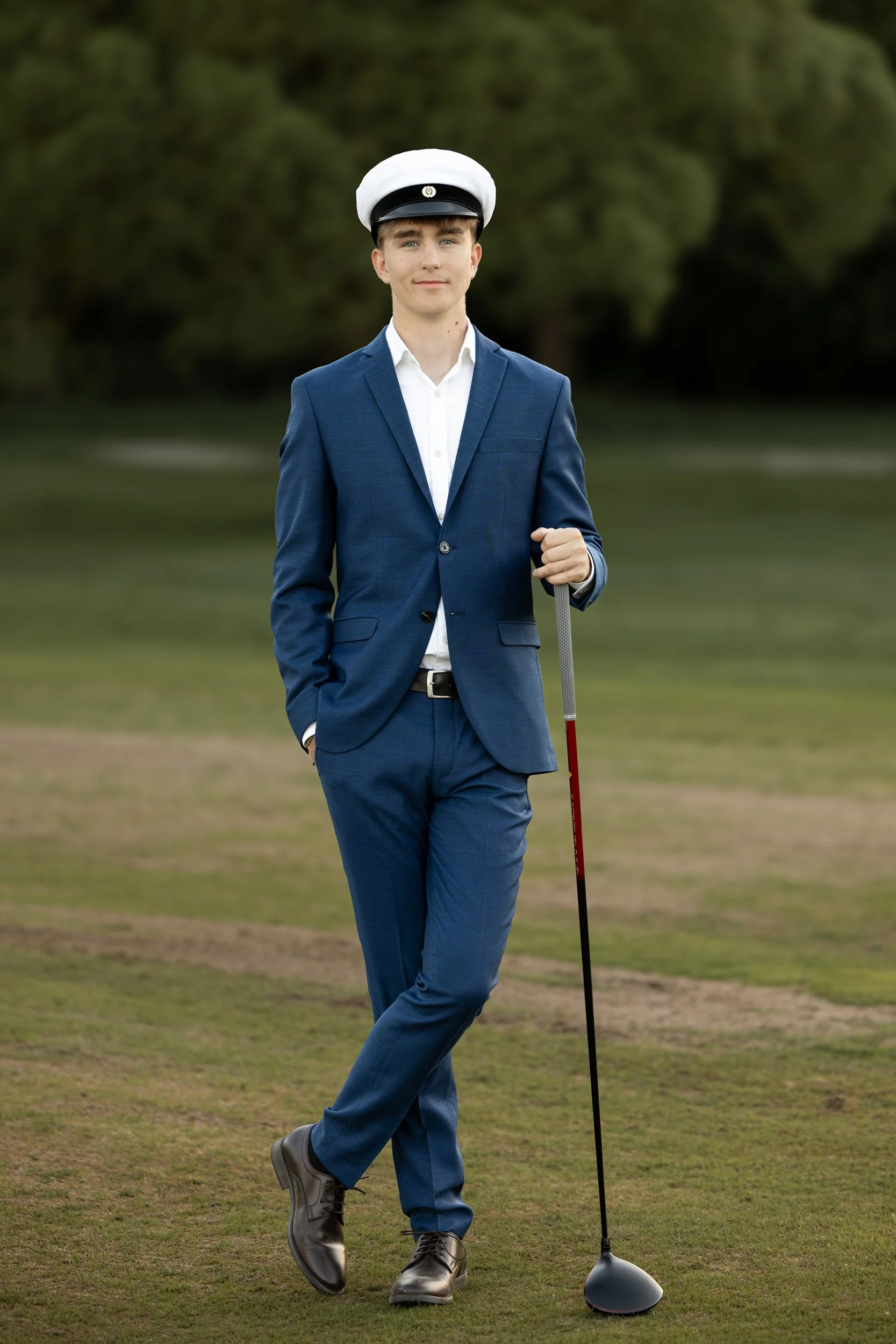 Young man in a blue suit with a white shirt and black shoes standing on a golf course, holding a golf club.