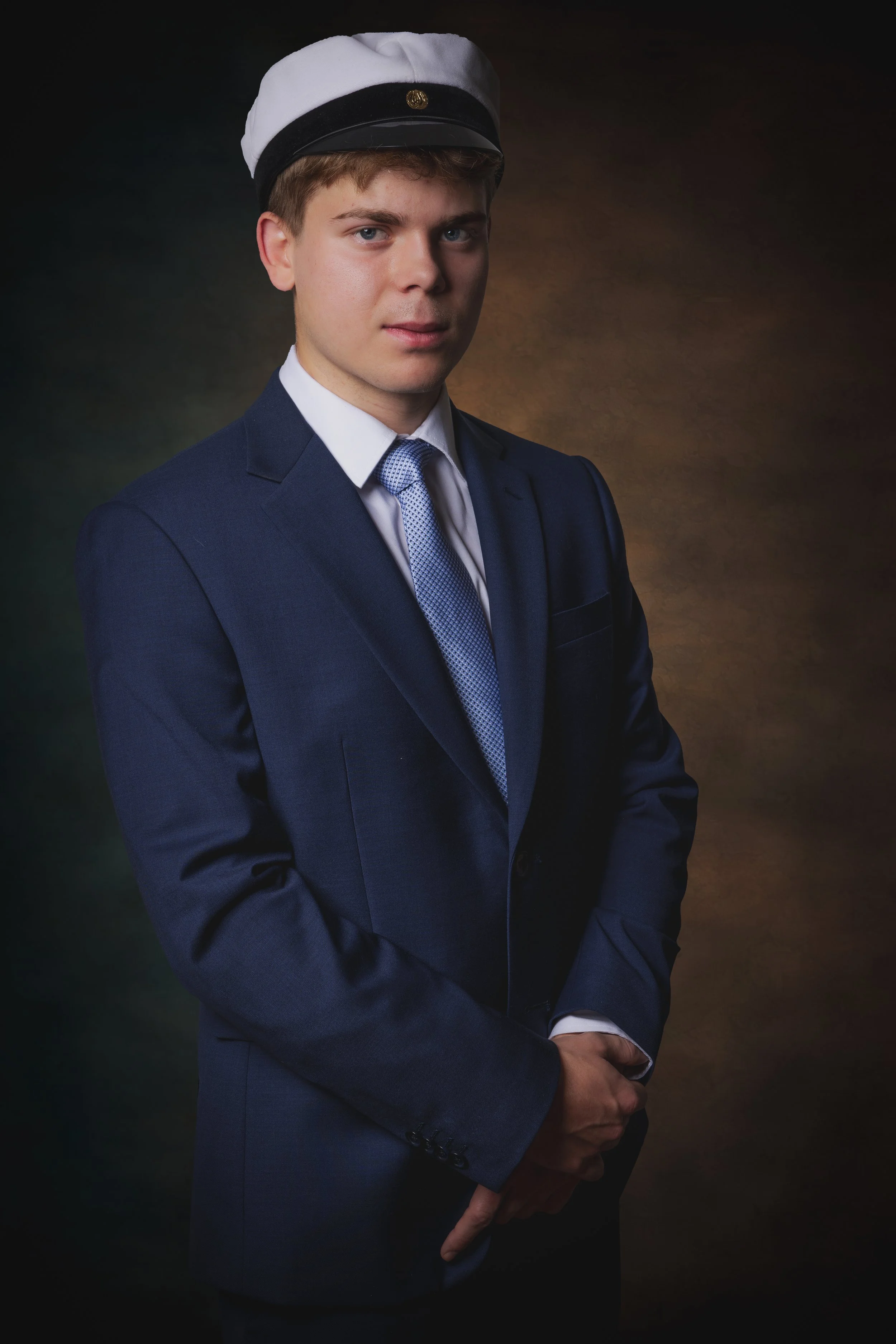 Young man in a navy blue suit and tie, wearing a white and black graduation cap, standing against a brown and dark background.