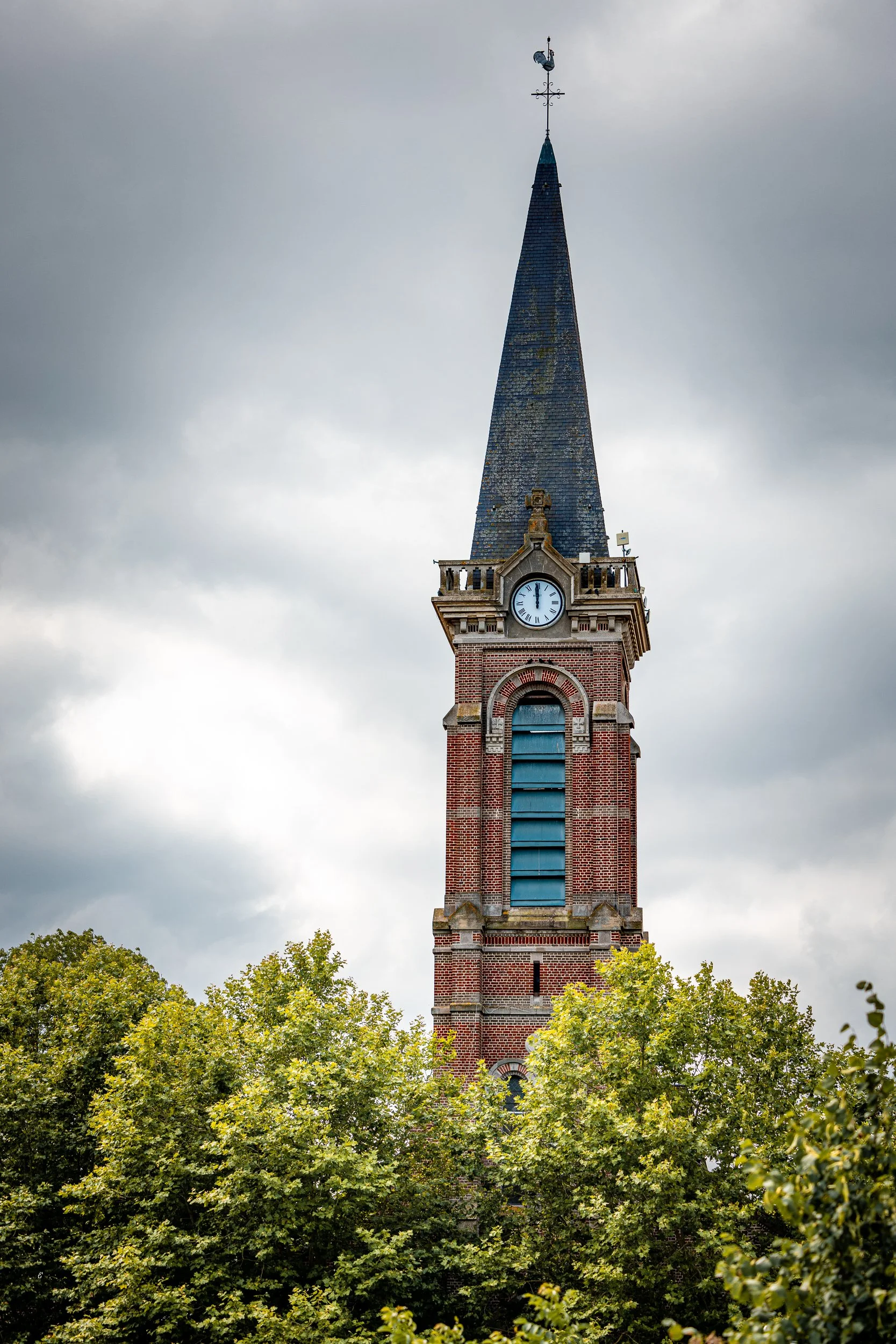 A tall church steeple with a clock near the top, partly obscured by green trees, under a cloudy sky.