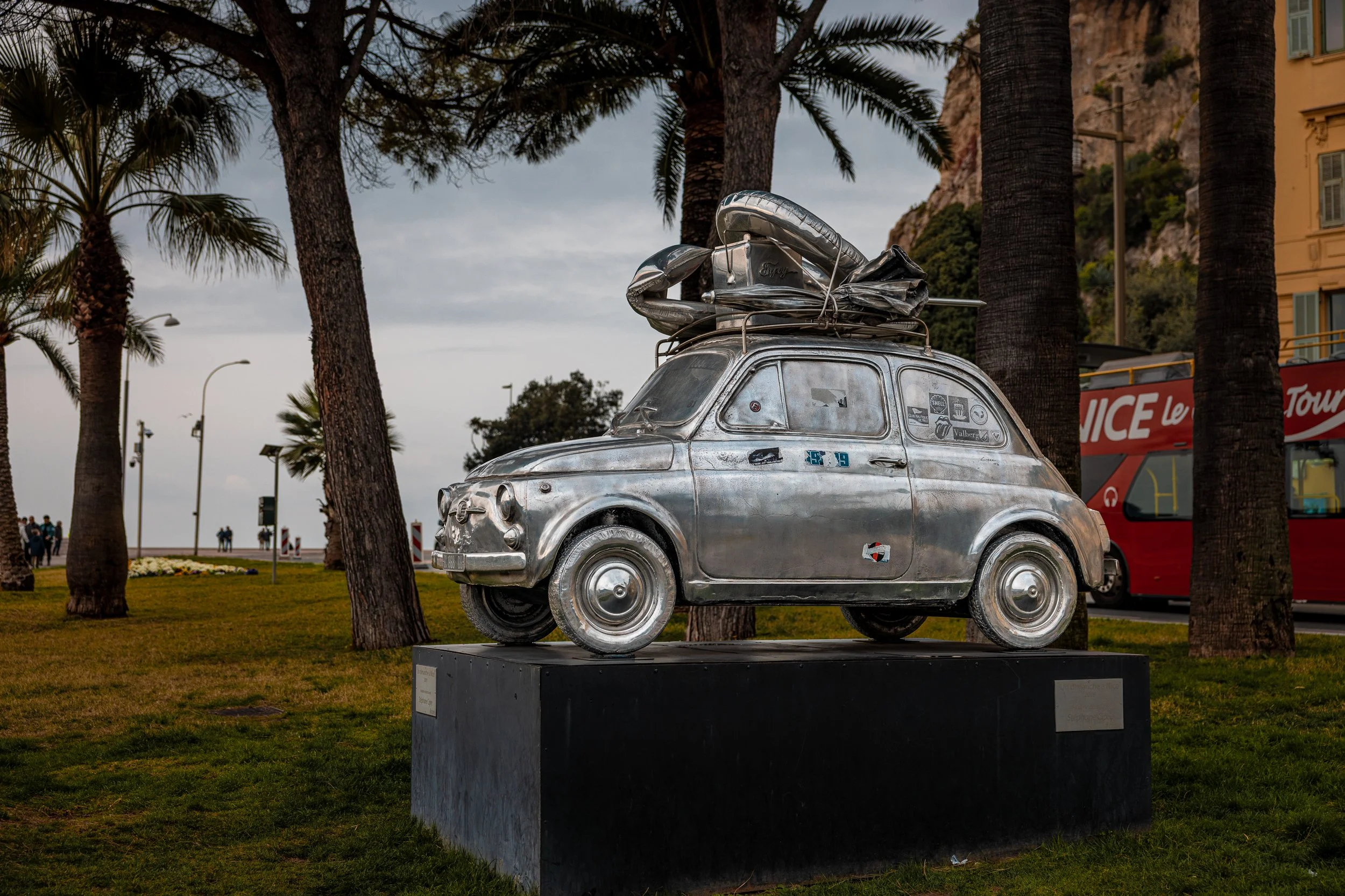 A metallic sculpture of a small vintage car with luggage on top, mounted on a black pedestal, situated in a park with palm trees, near a coastline, with a tour bus in the background.