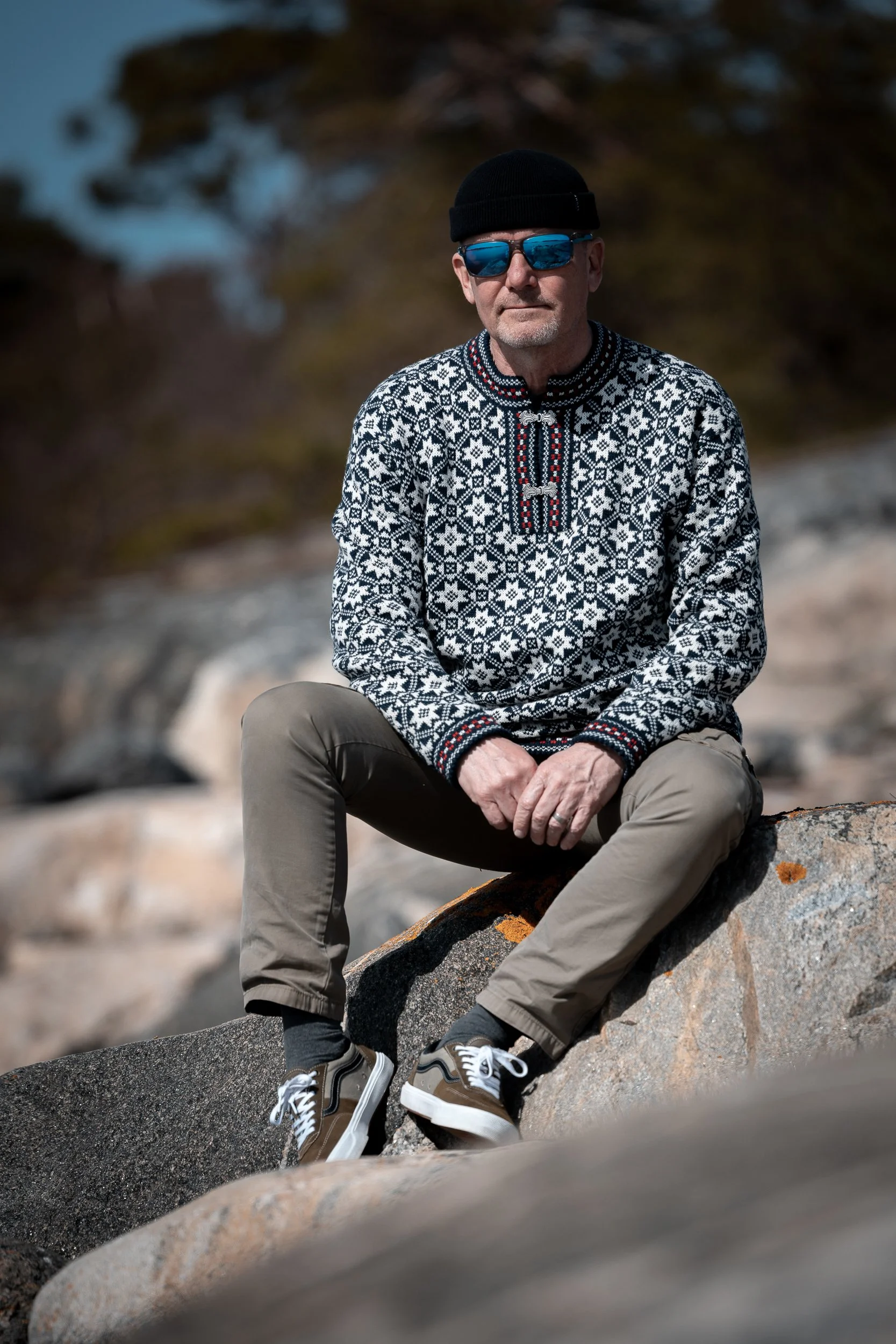 A man sitting on a rock outdoors wearing sunglasses, a beanie, a patterned sweater, khaki pants, and sneakers.