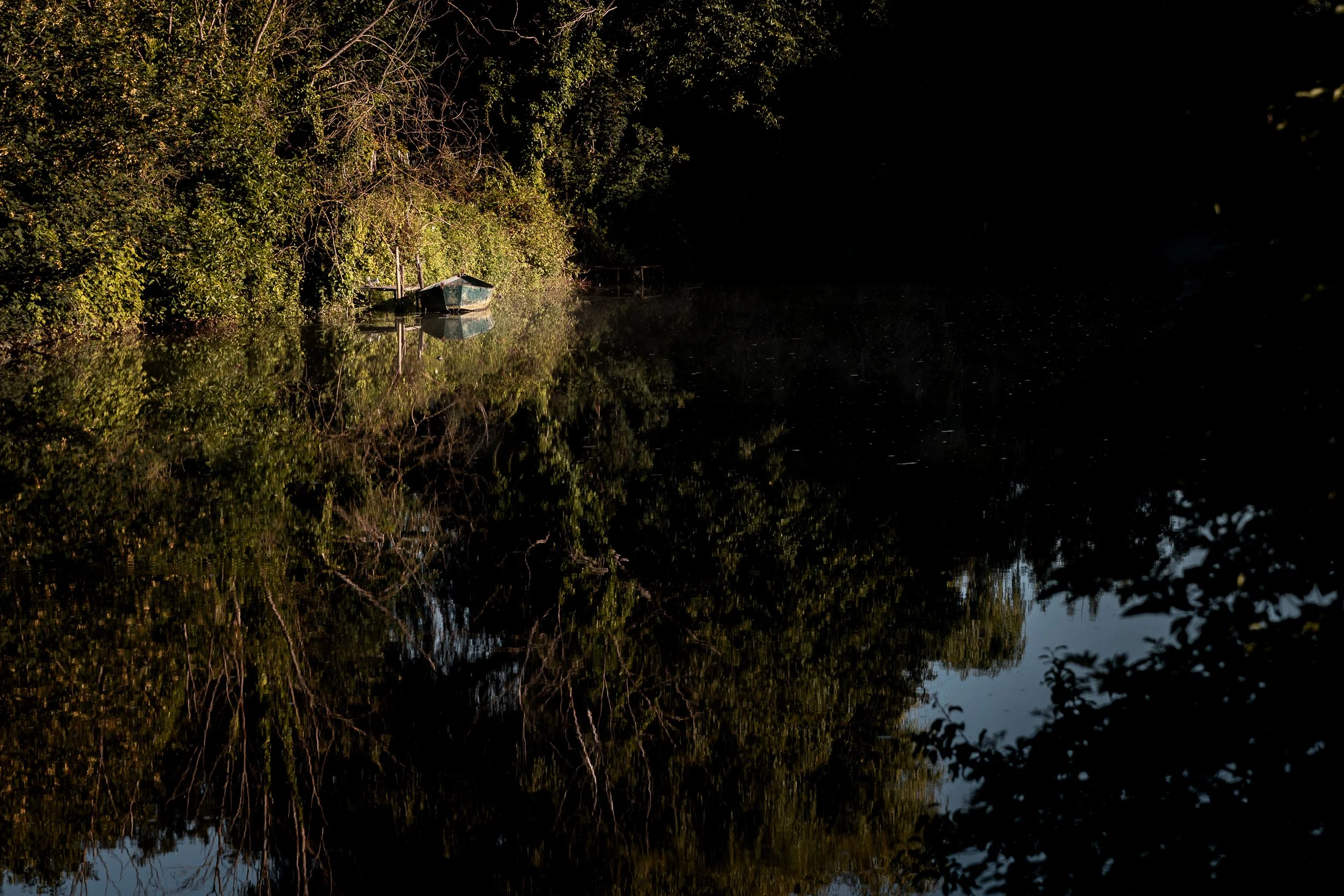 A small boat is floating on a calm body of water, surrounded by lush green trees reflected in the water, with shadows indicating it is either early morning or late afternoon.