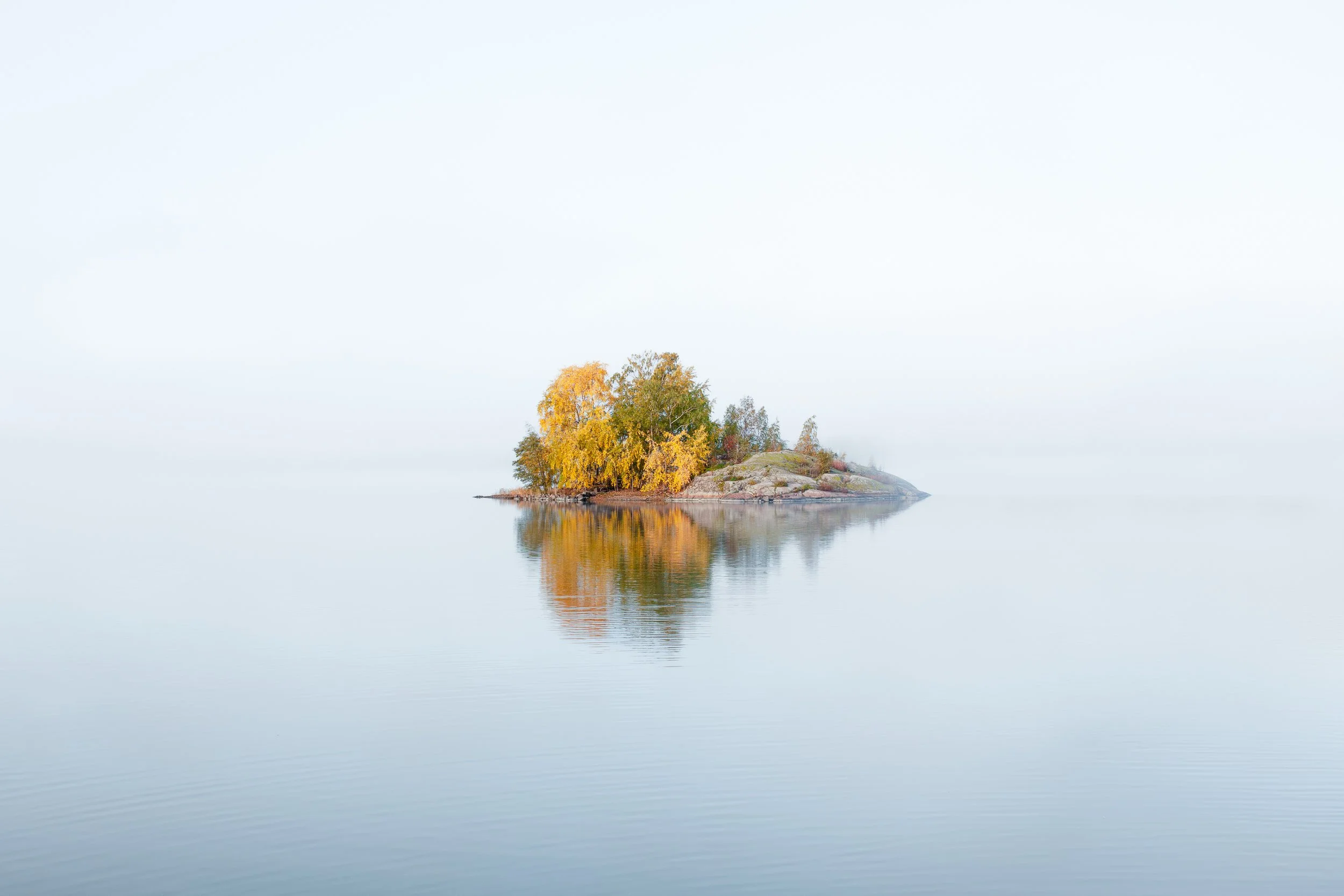 A small island with trees displaying autumn colors reflected in calm water.