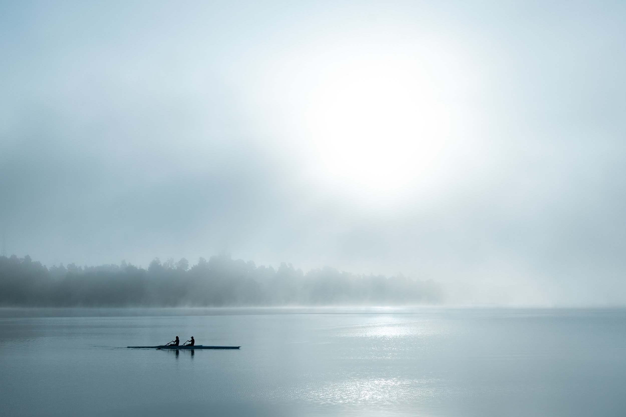Two people rowing a boat on a calm, misty lake with a tree-lined shoreline in the background, under a bright, cloudy sky.