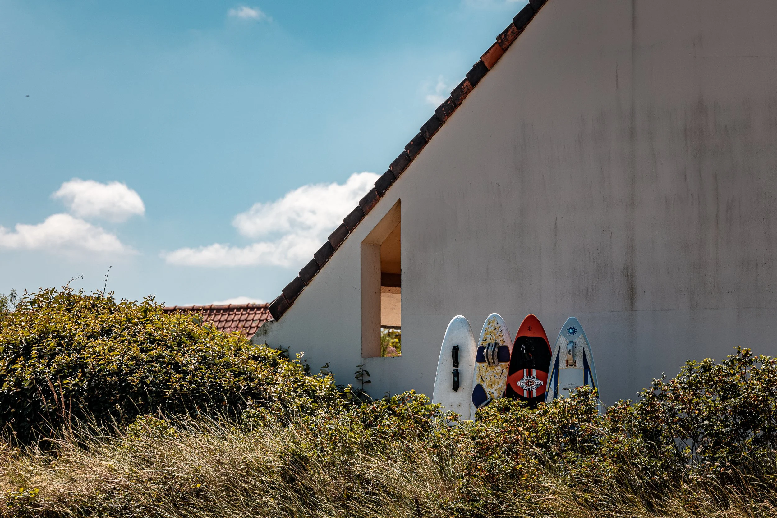 Four stand-up paddleboards leaning against a white wall of a building with a red-tiled roof, surrounded by bushes and tall grass, under a blue sky with scattered clouds.