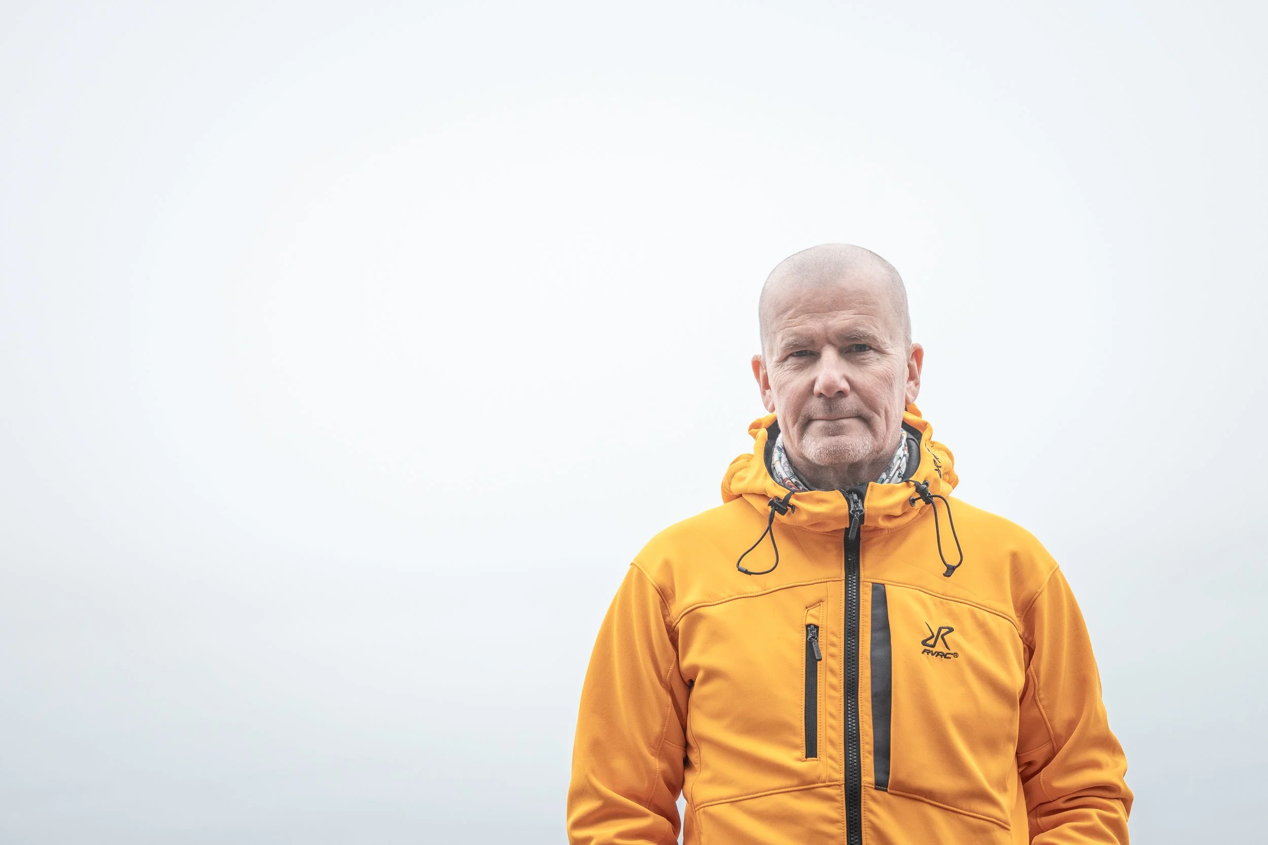 A man with a serious expression wearing a bright orange outdoor jacket against a plain, overcast sky.