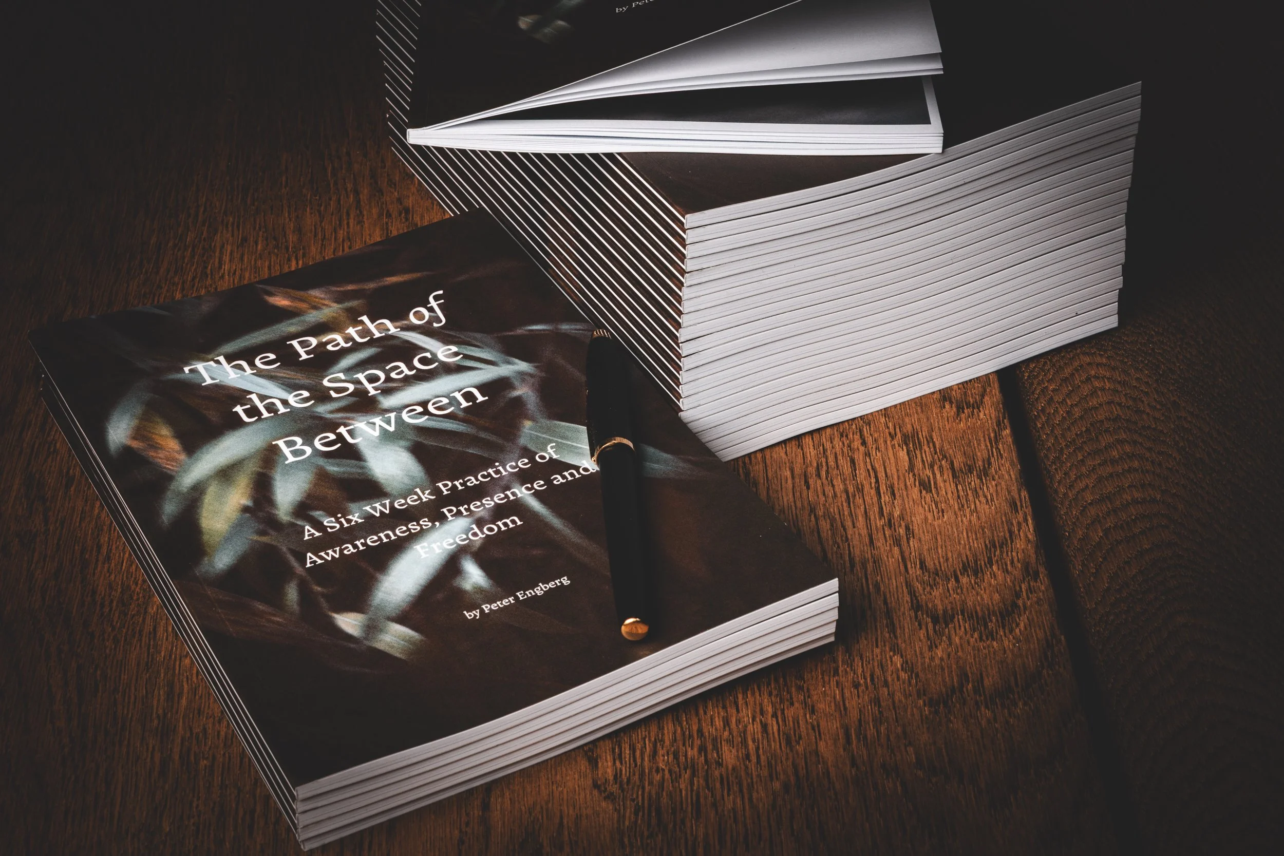 Book titled 'The Path of the Space Between' by Peter Engberg, placed on a wooden surface with a black and gold pen on top, next to a large stack of similar books.