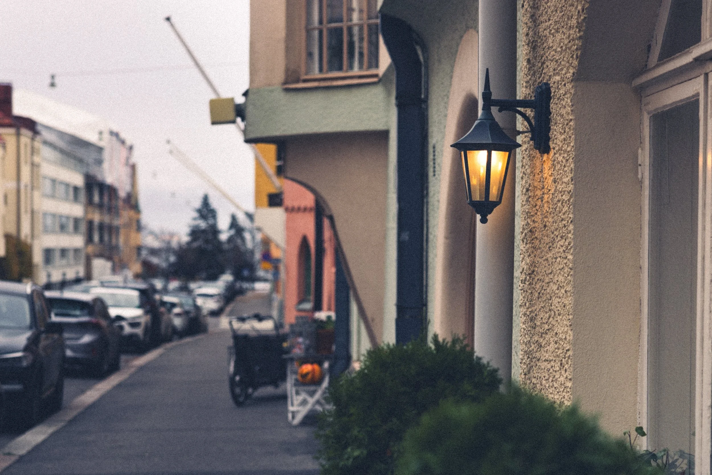 Street view with colorful houses, parked cars, and a lit outdoor wall lamp during cloudy evening.