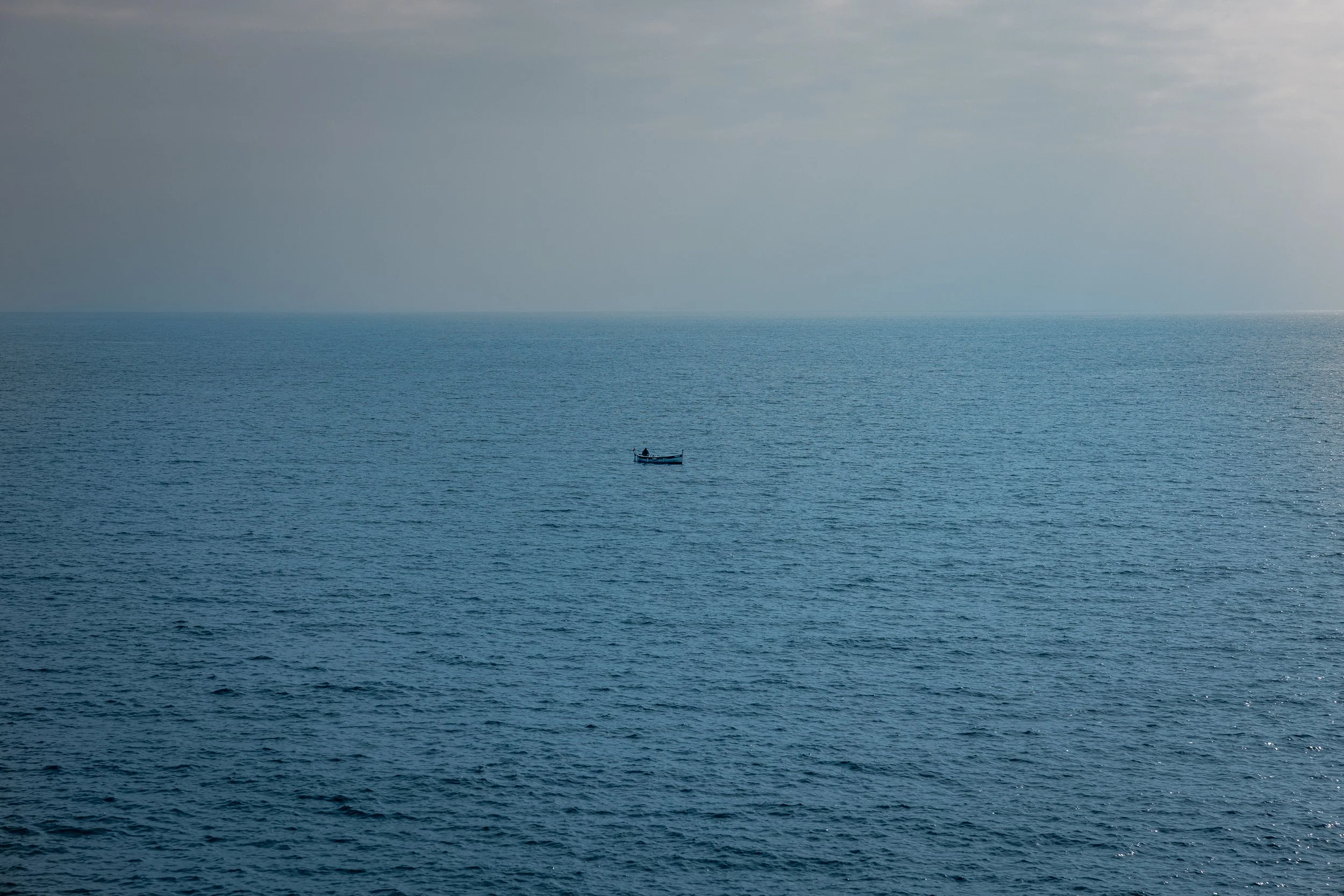 A small boat floating in a calm, expansive ocean under a cloudy sky.