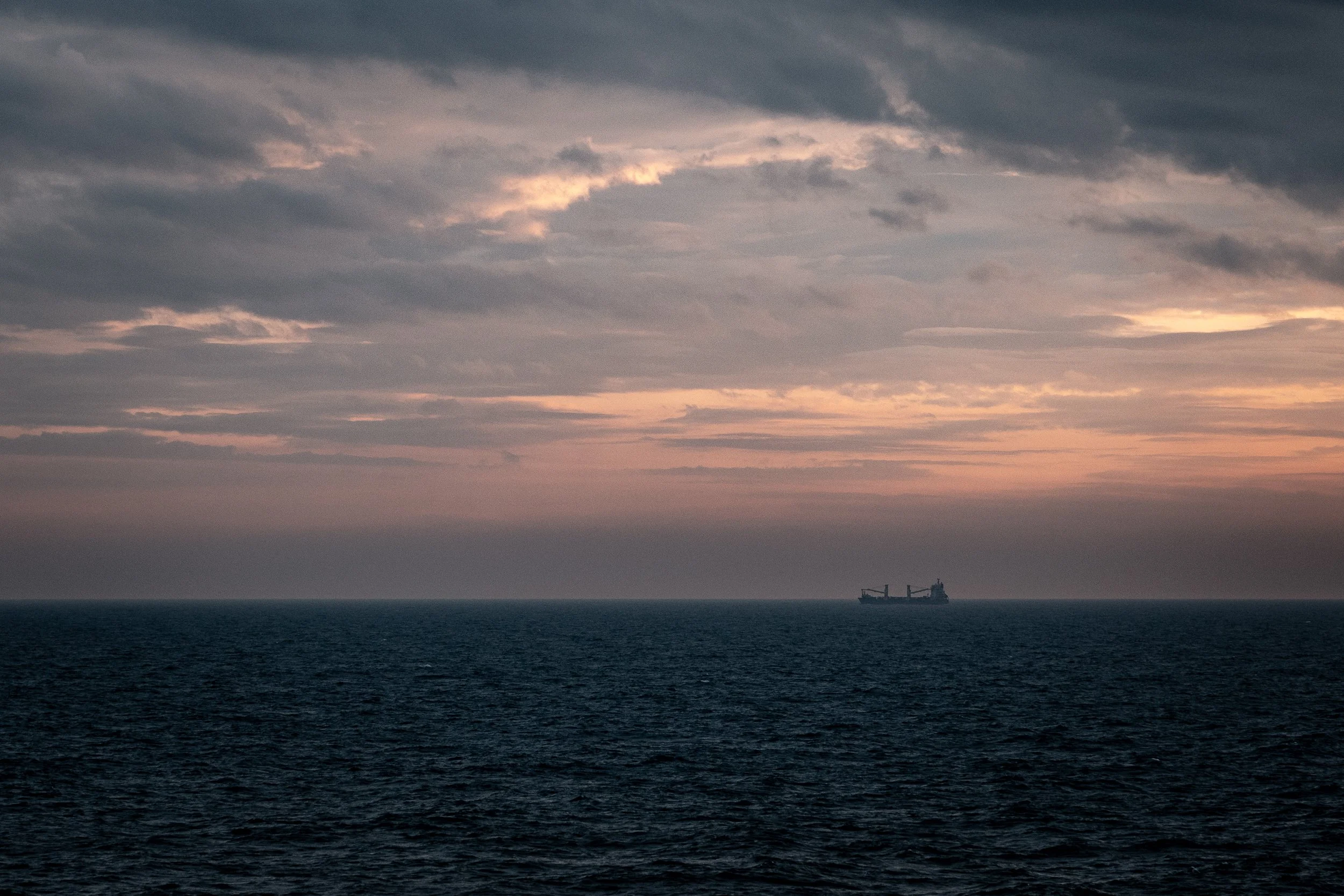 A large cargo ship is on the horizon in a vast ocean at sunset, with a sky filled with dark clouds and patches of orange and pink light.