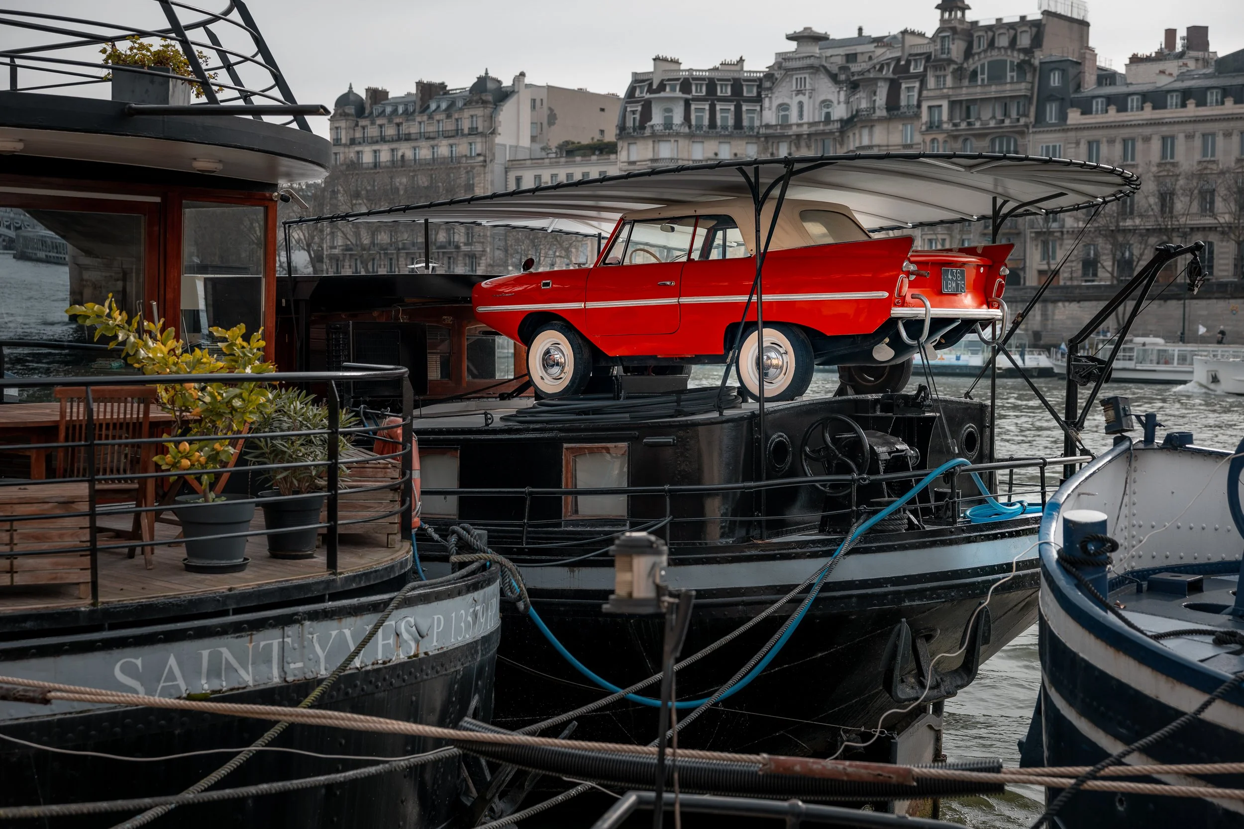 A red vintage car mounted on the roof of a black boat docked on a river, with old buildings in the background.