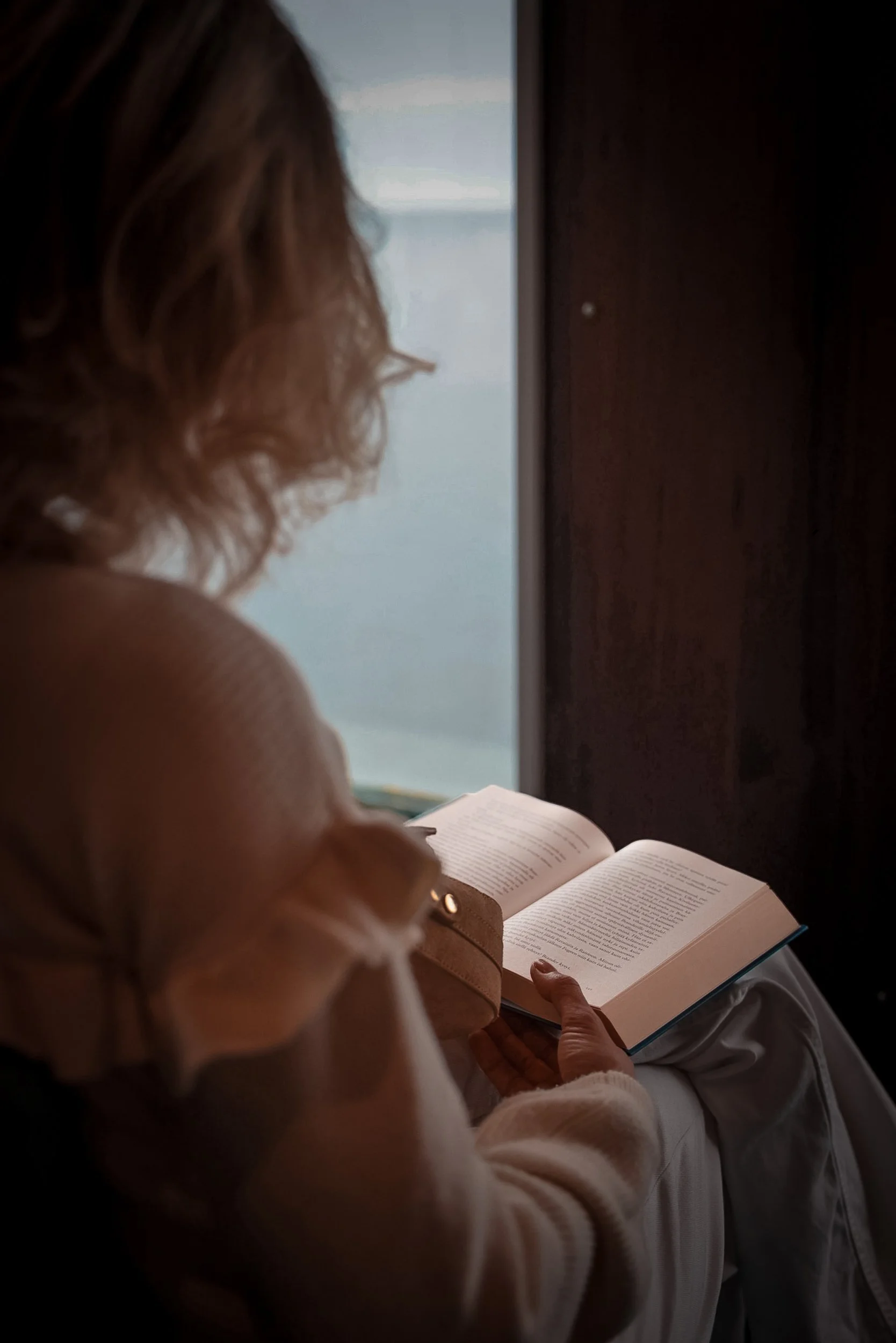A woman with curly hair reading a book on her lap while sitting by a window.