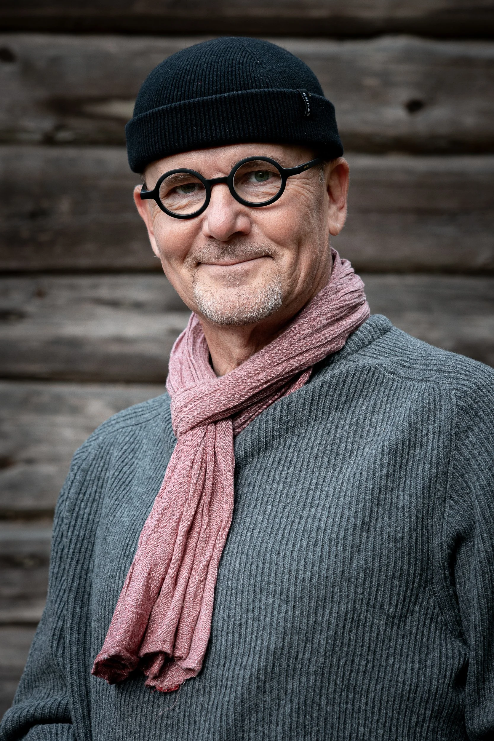 A middle-aged man wearing a black knit hat, round black glasses, a pink scarf, and a gray sweater, standing in front of a wooden background.