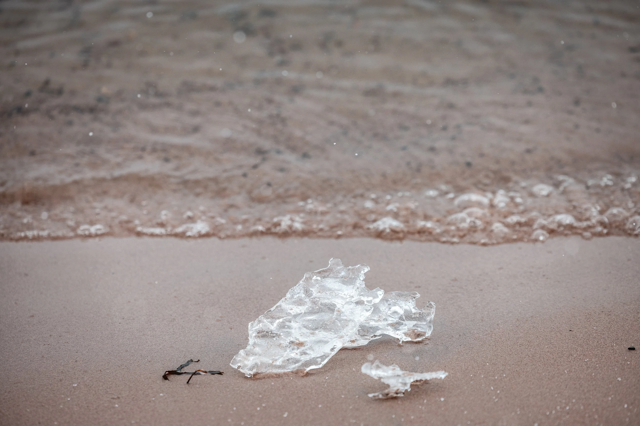 A piece of clear, broken ice on sandy beach with small piece nearby and ocean waves approaching in the background.