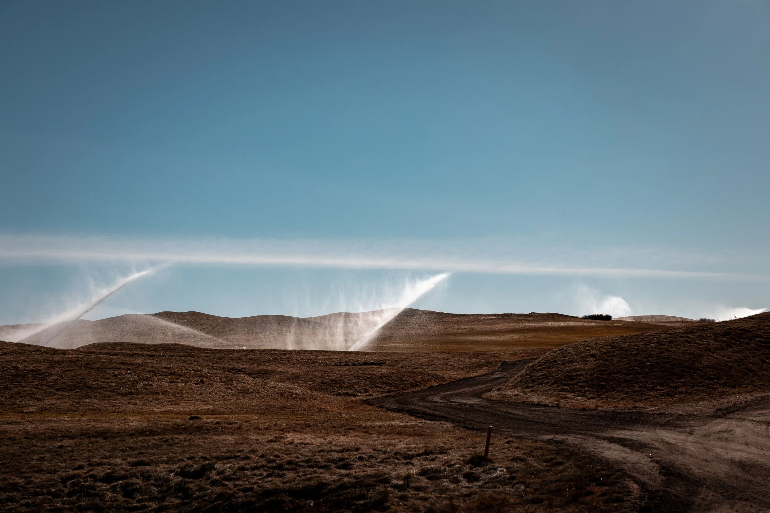 Dirt road winding through dry, rolling brown hills under a clear blue sky with water sprays in the distance.