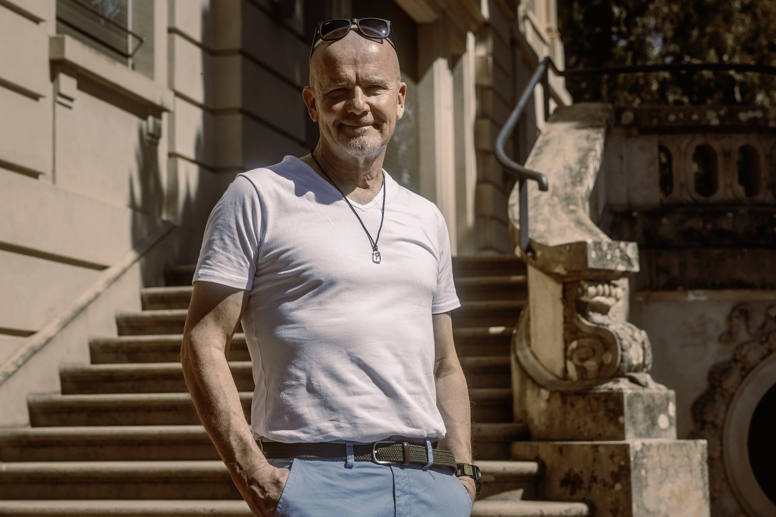 A ikääntynyt mies hymyilee ulkona, hänellä on päällään valkoinen t-paita ja vaaleansiniset housut. Taustalla on vanha kivitalo ja kiviterassi, jossa on kaide ja portaat.