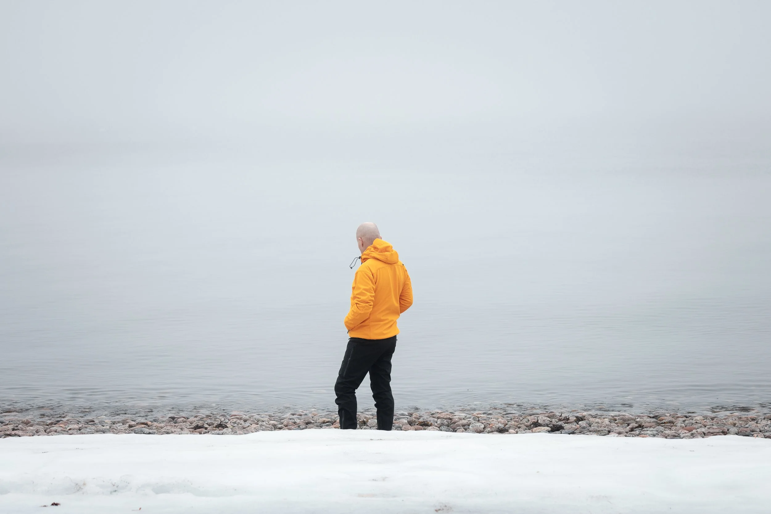A person with a shaved head wearing a yellow jacket and black pants standing on a rocky beach near calm water, facing away from the camera.