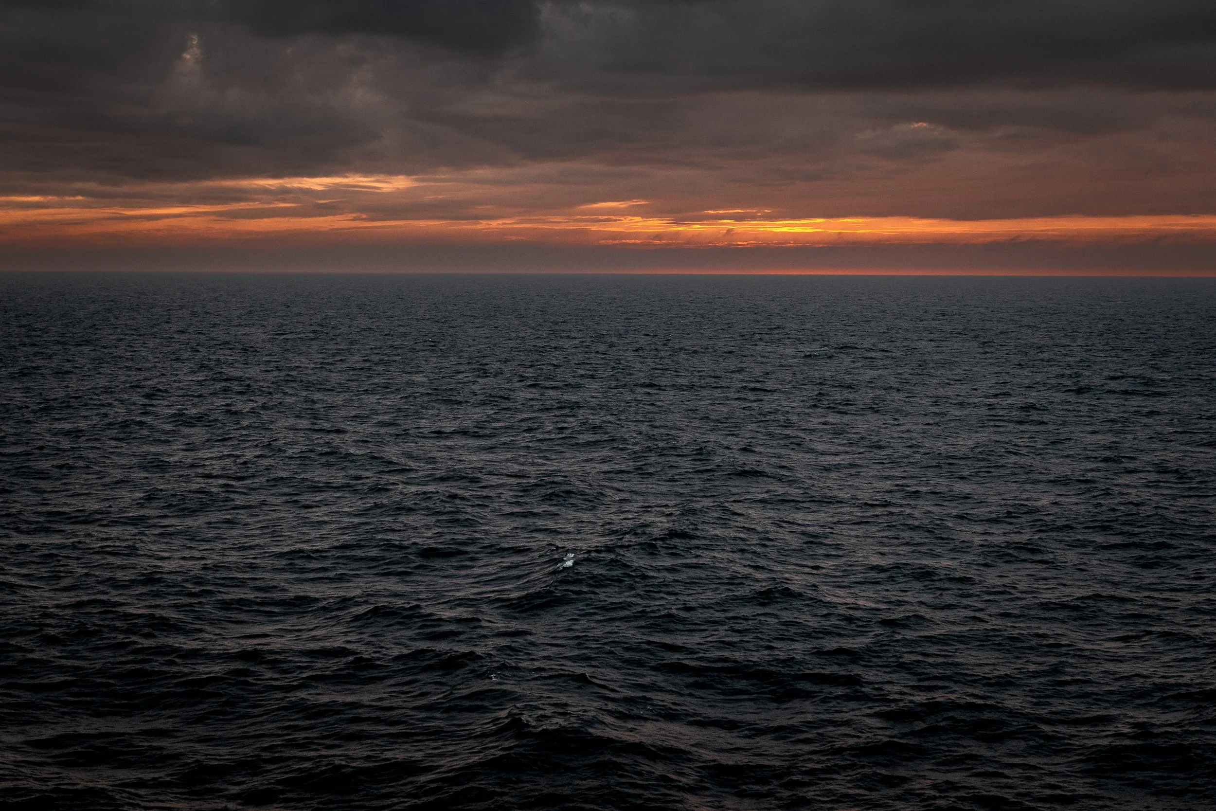 A vast ocean with dark, choppy waters under a cloudy sky during sunset or sunrise, with a hint of orange and yellow near the horizon.