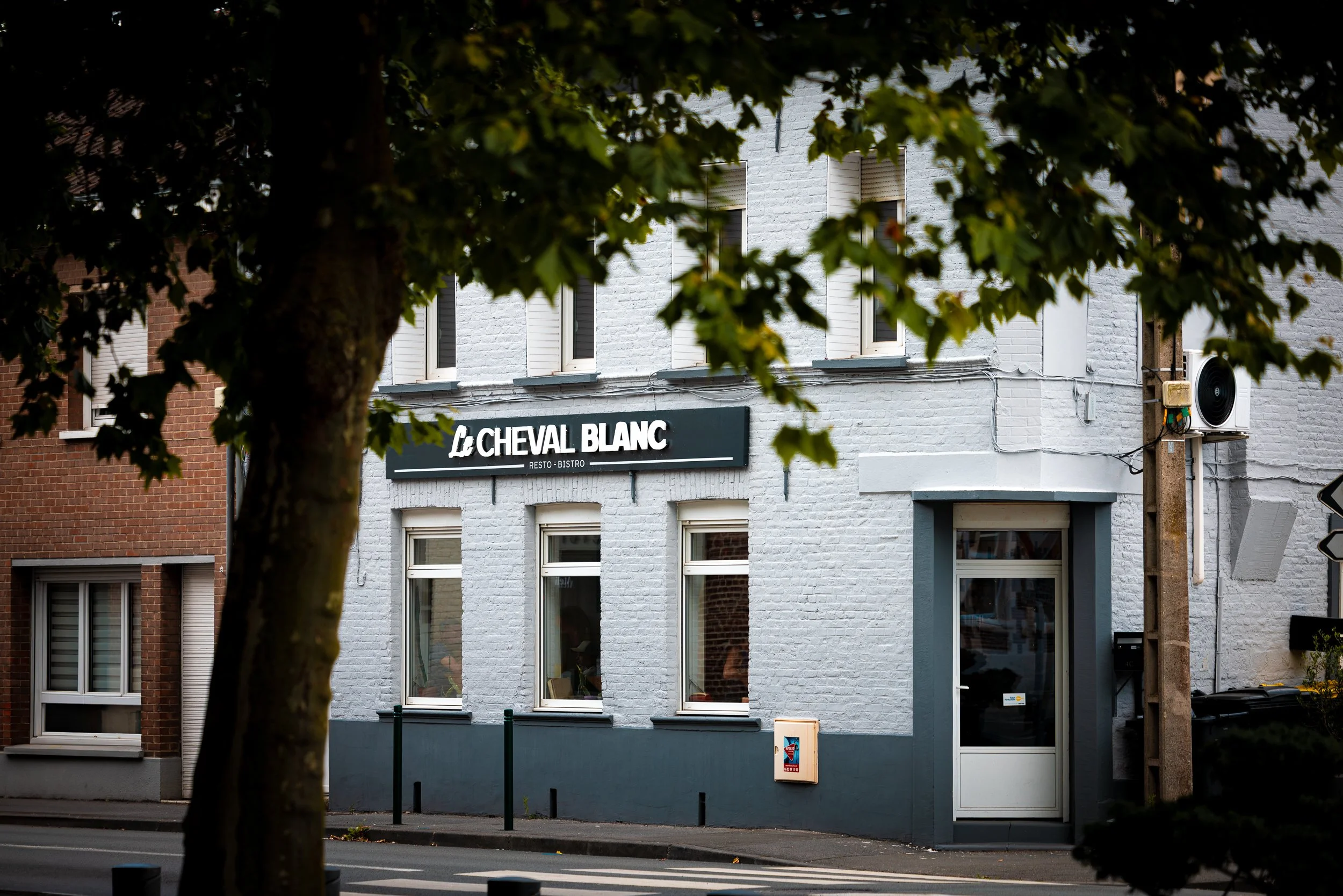 A white brick building with a sign that reads 'Le Cheval Blanc Resto - Bistro' and shows a street view with a tree in the foreground.