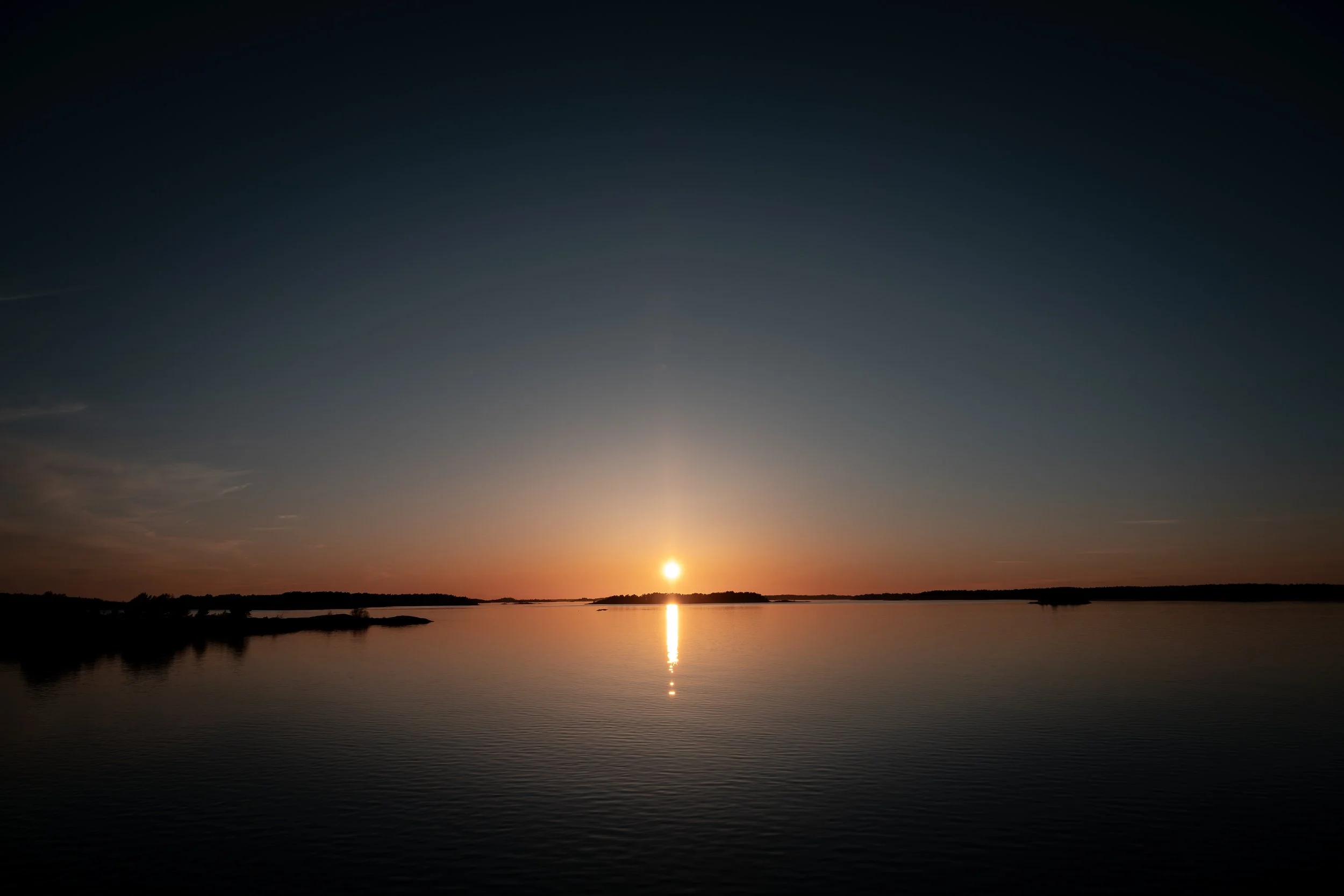 Sunset over a calm body of water with a reflection of the sun on the surface, mostly clear sky with a few clouds.
