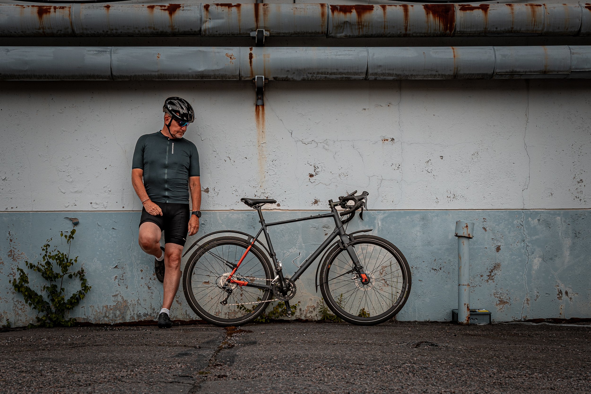 A man wearing a bike helmet and cycling clothes standing against a weathered wall with his bike, stretching his leg on the lowest pedal of the bicycle.