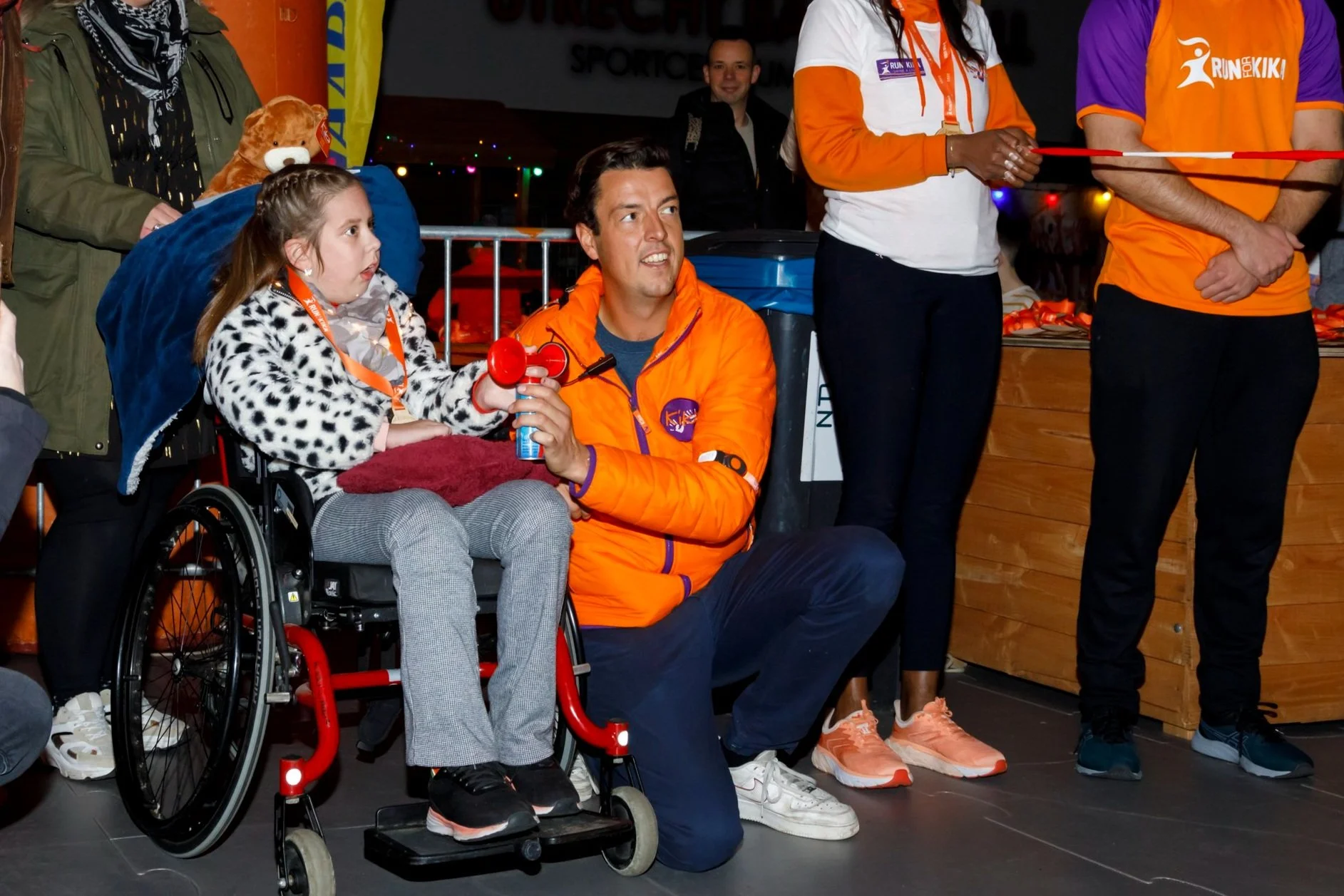 Rick en een meisje in een rolstoel die deel uitmaken van een evenement voor KiKa, omringd door mensen, met vriendelijke sfeer en kleurrijke kleding om geld op te halen tegen kanker.