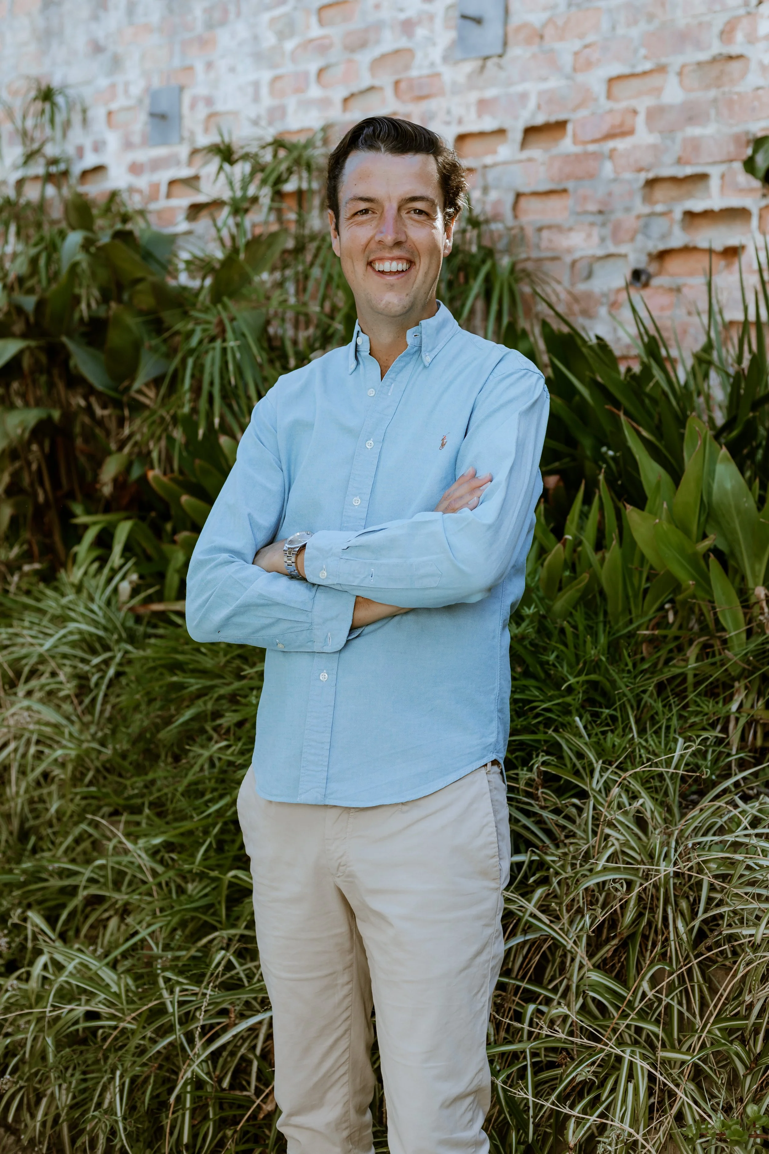 Man met donkere haar in een lichte blauwe overhemd en beige broek, glimlachend, staand voor groen gebladerte en een bakstenen muur.