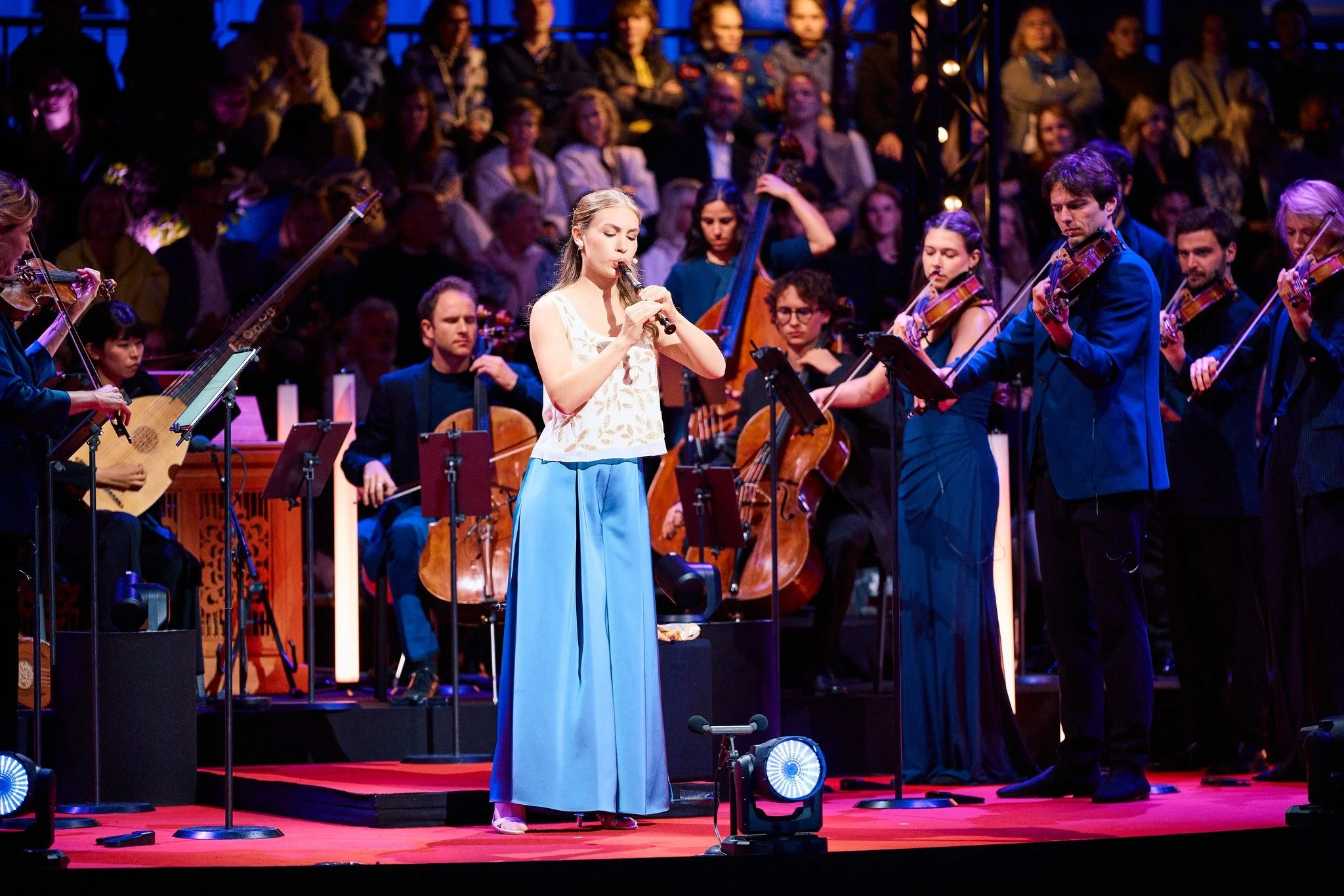 Symfonieorkest met een vrouw die fluit speelt op een podium met een grote publiek op de achtergrond tijdens Prinsengrachtconcert.