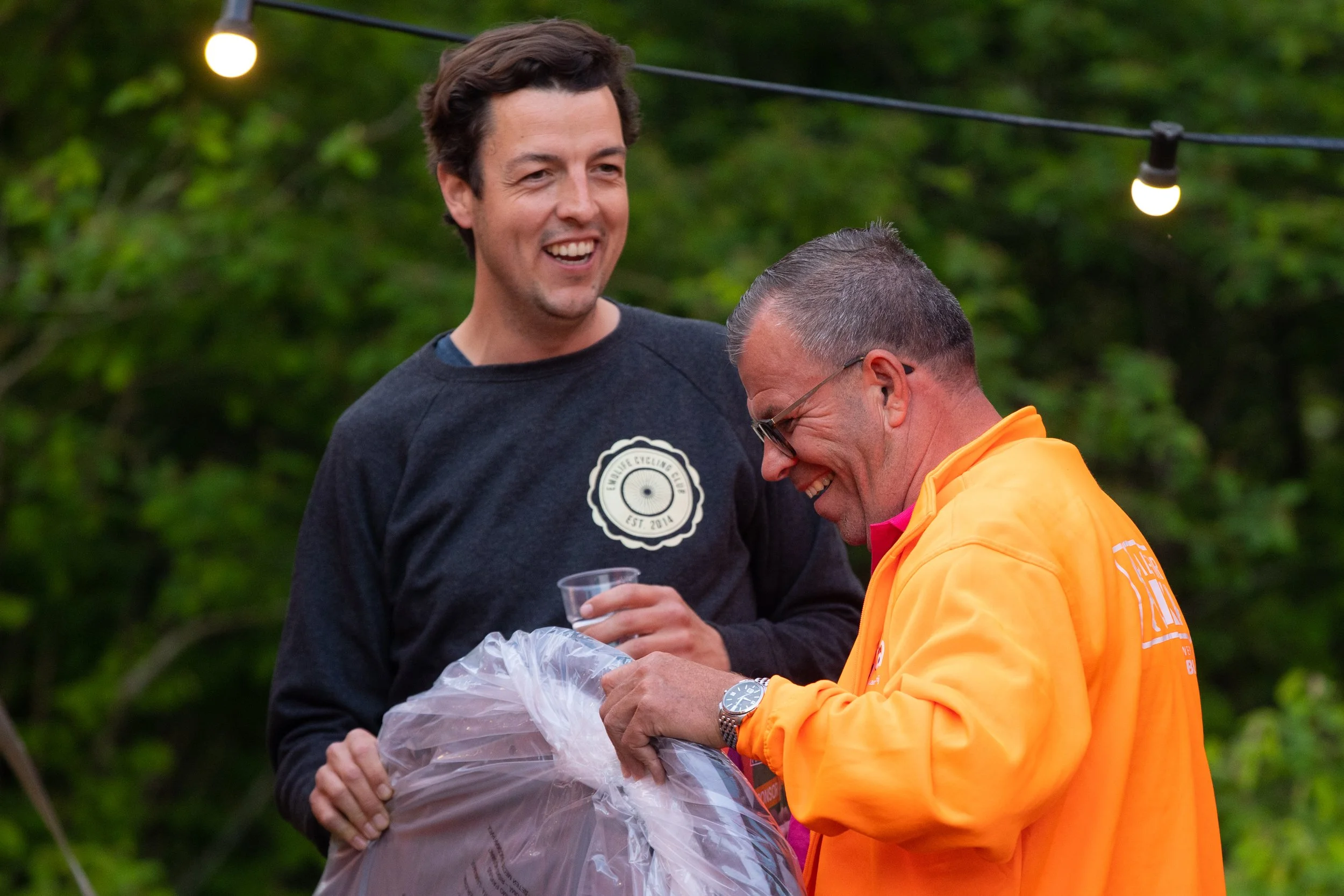 Twee mannen lachen en praten tijdens een buitenbijeenkomst, één in een zwart shirt en de andere in een oranje jas, onder stringverlichting met groene bomen op de achtergrond.