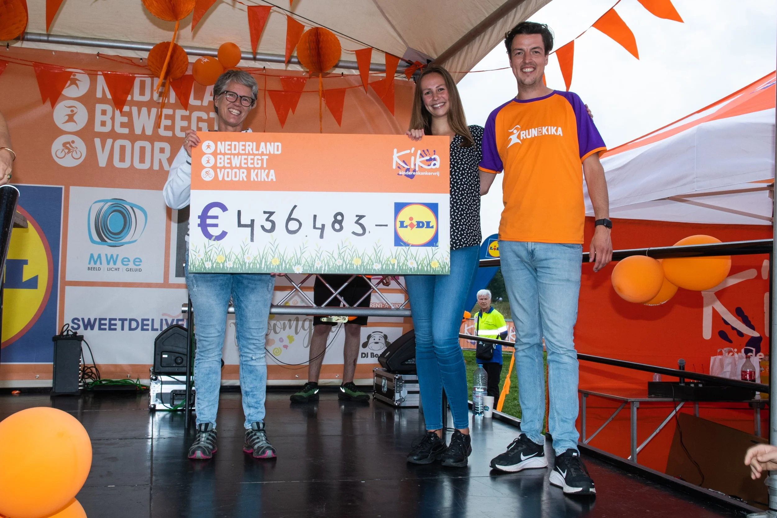 Drie mensen staan op een podium, vieren een cheque-uitreiking met een grote cheque van €436.483, in Utrecht, tijdens een evenement, met oranje decoraties en banners voor KIKA en Lidl.