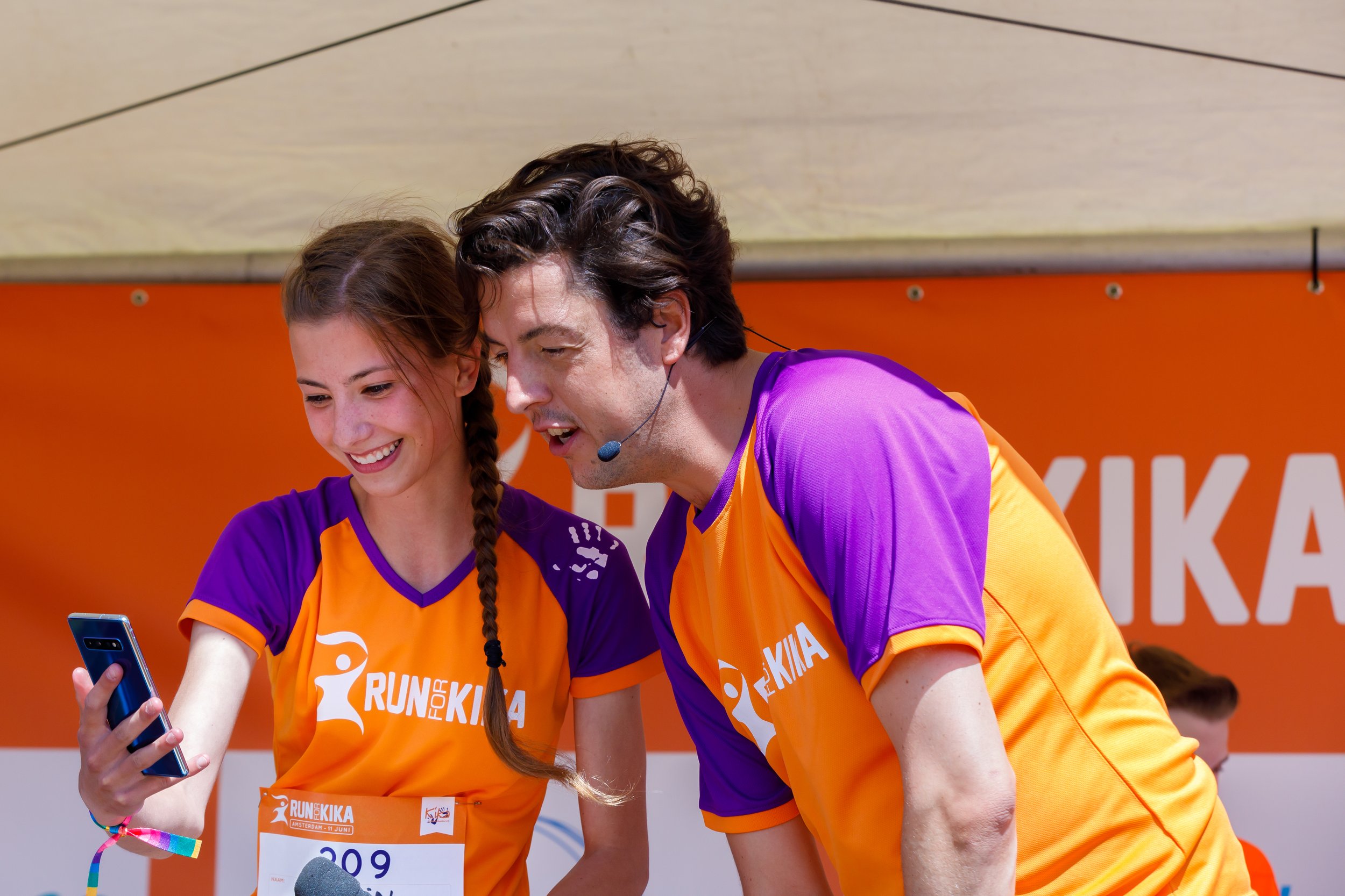 Twee lopers in oranje en paarse shirts bekijken een smartphone samen, ze lachen en zijn op een evenement met een oranje achtergrond.