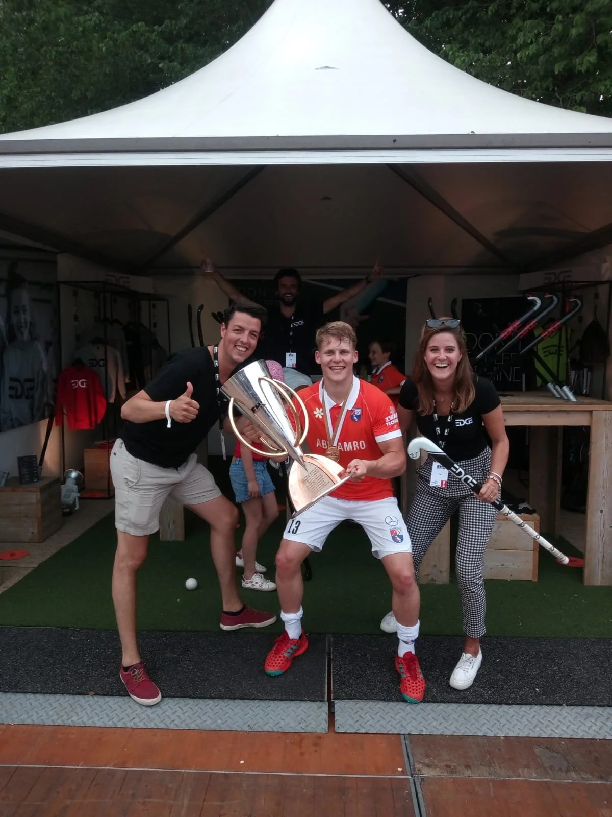 Speler van hockeyclub Bloemendaal met beker, omringd door Rick en Esmee. Ze vieren de overwinning van Bloemendaal tijdens de Euro Hockey League (EHL) en staan voor de winkel van EDGE, het hockeymerk van Rick.