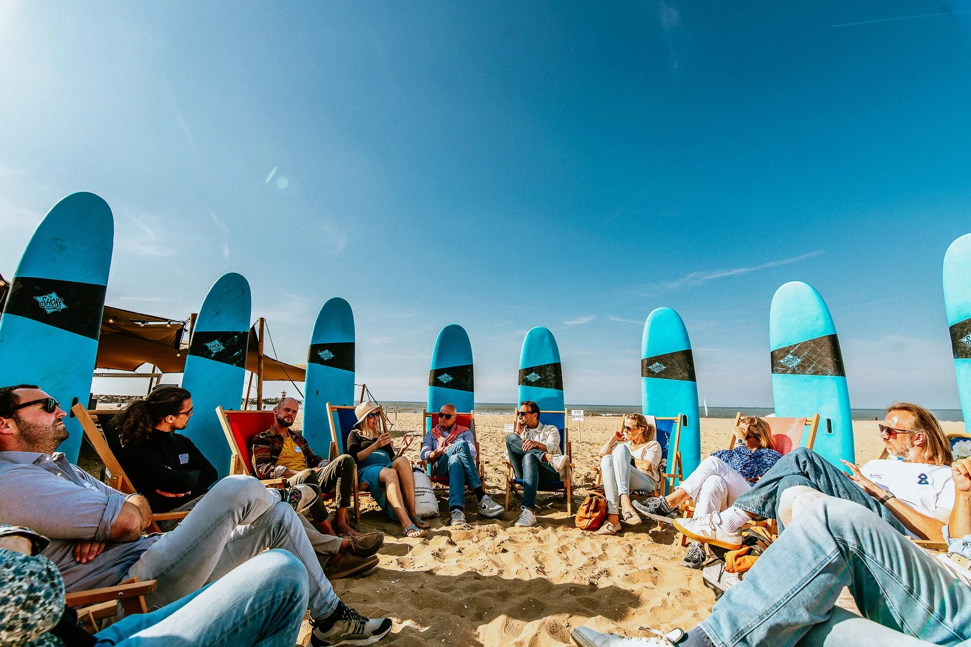 Mensen zitten op strandstoelen onder een blauwe parasol, omringd door blauwe surfplanken, op een zandstrand met een heldere blauwe lucht.