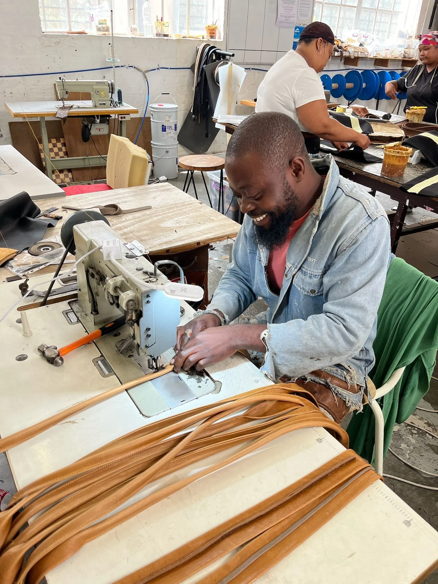 Man die met een naaimachine leerachtige riemen maakt in een atelier van Rowdy uit Zuid Afrika, met andere mensen en materialen op de achtergrond.