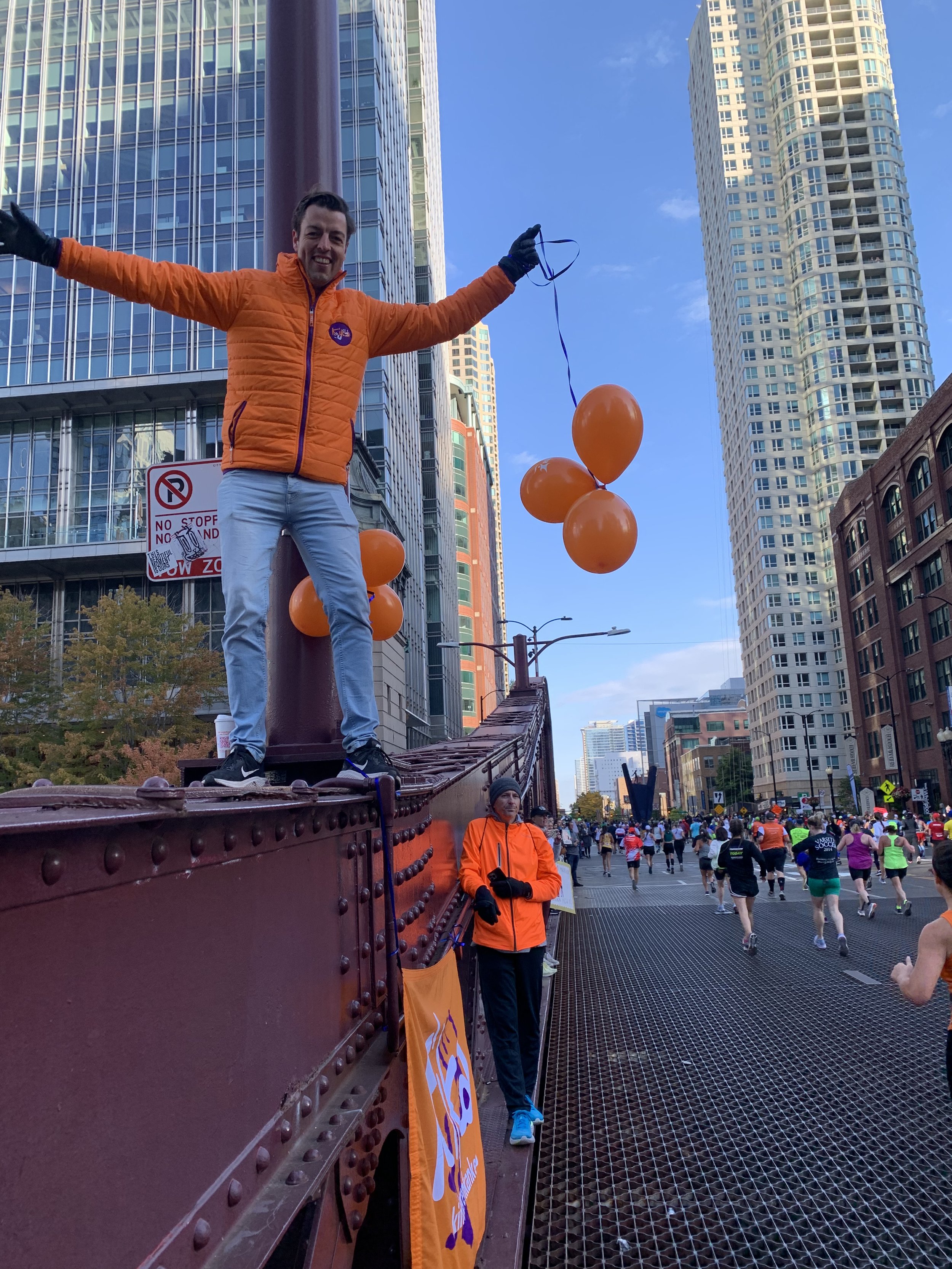 Een man staat op een brug in Chicago tijdens de marathon en zwaait met zijn armen uit terwijl hij drie oranje ballonnen vasthoudt. Er is ook een vrouw in een oranje jas die onder de brug staat, en samen moedigen ze de lopers aan.