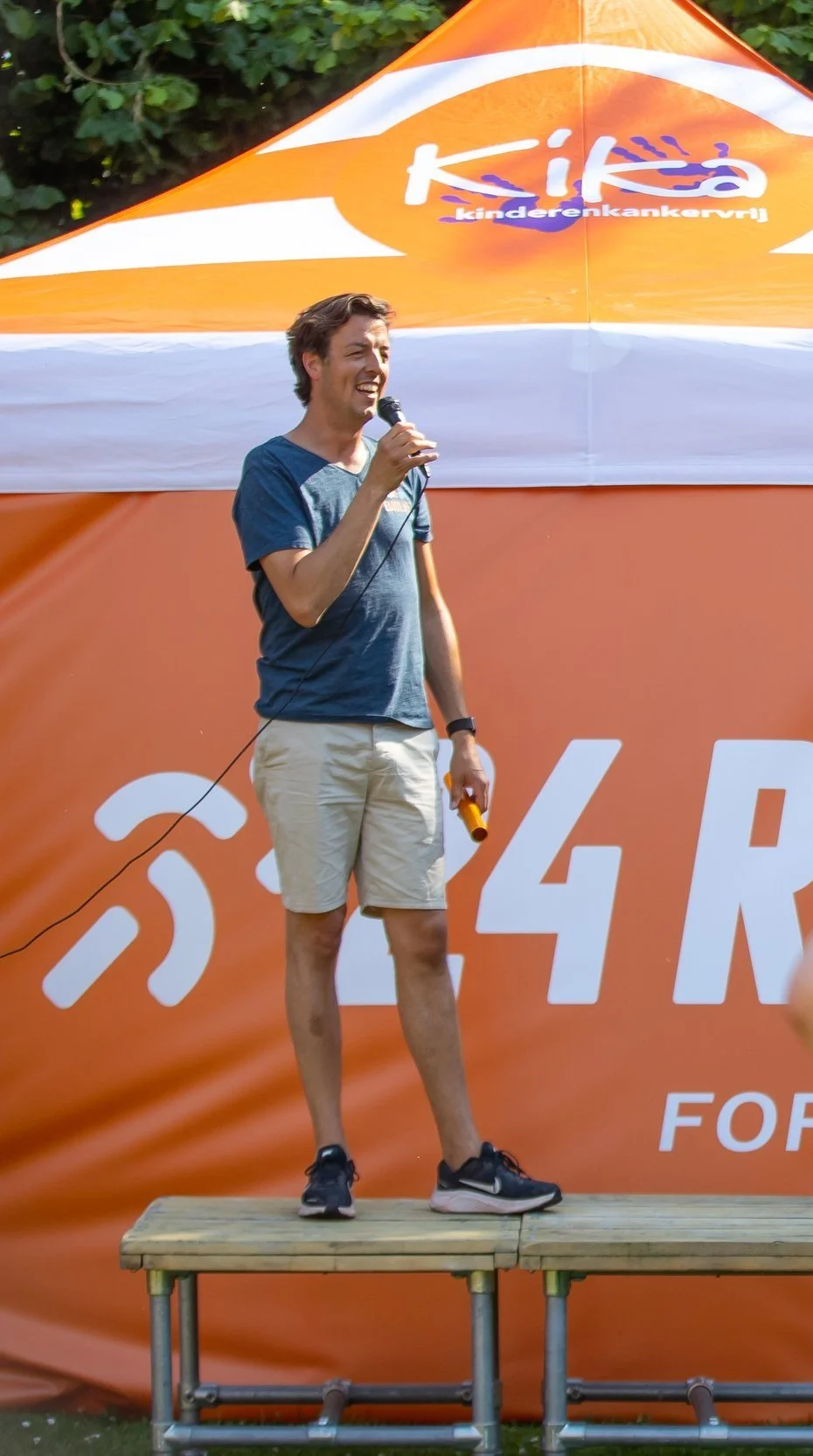 Man spreekt op een podium met een microfoon in zijn hand, voor een oranje en witte tent met het logo 'KiKa' en de tekst 'kinderenkankervrij' eronder.