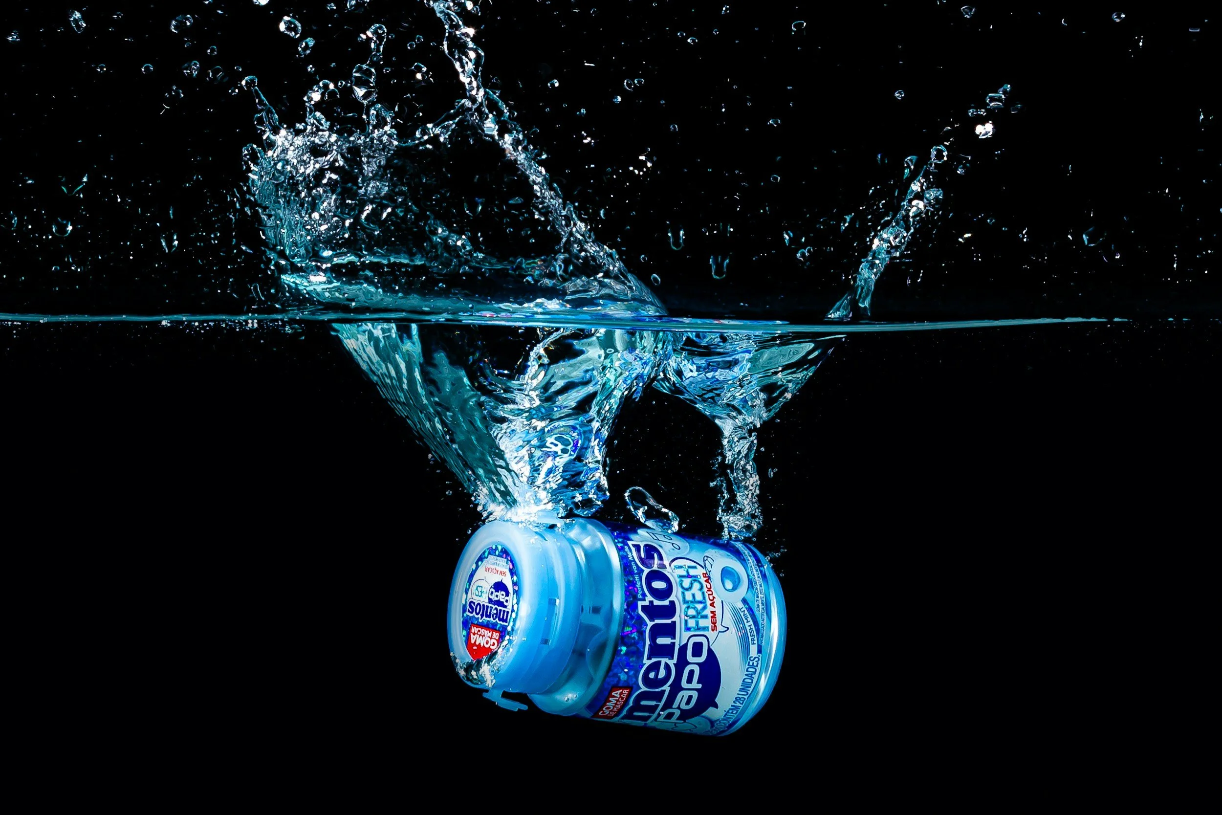 Ein Glas mit Wasserspritzern beim Eintauchen in Wasser, darunter eine Dose mit blauber Schrift auf schwarzem Hintergrund.