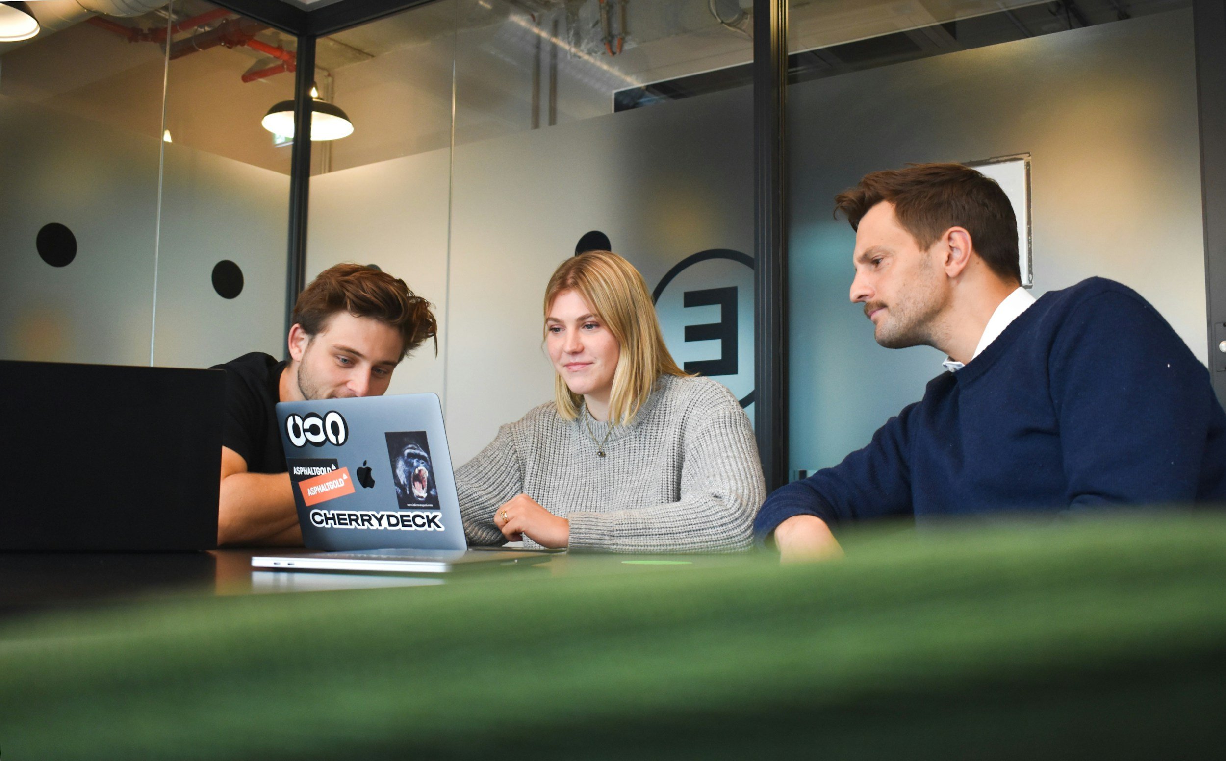 Drei junge Erwachsene sitzen an einem Tisch in einem modernen Büro, schauen auf einen Laptop und diskutieren, während sie sich auf die Arbeit konzentrieren.