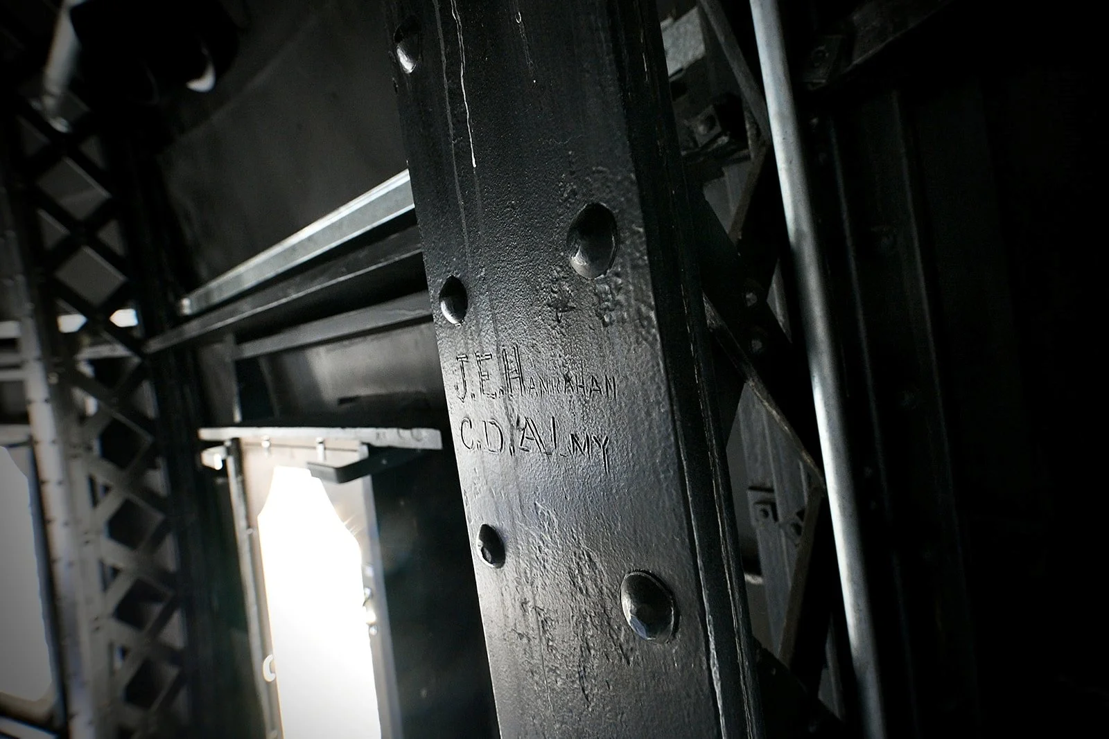 close up image of a metal truss with several names engraved into the metalwork, with more trusses and a window in the background.