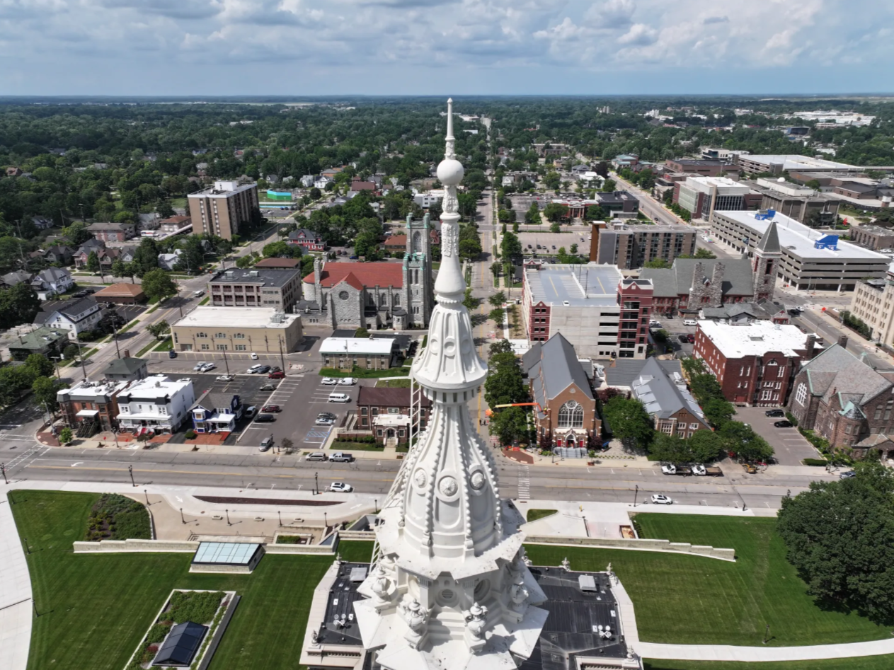 view of the top of the capitol, showing a tall, thin spire, with buildings and streets and trees in the distance below it.