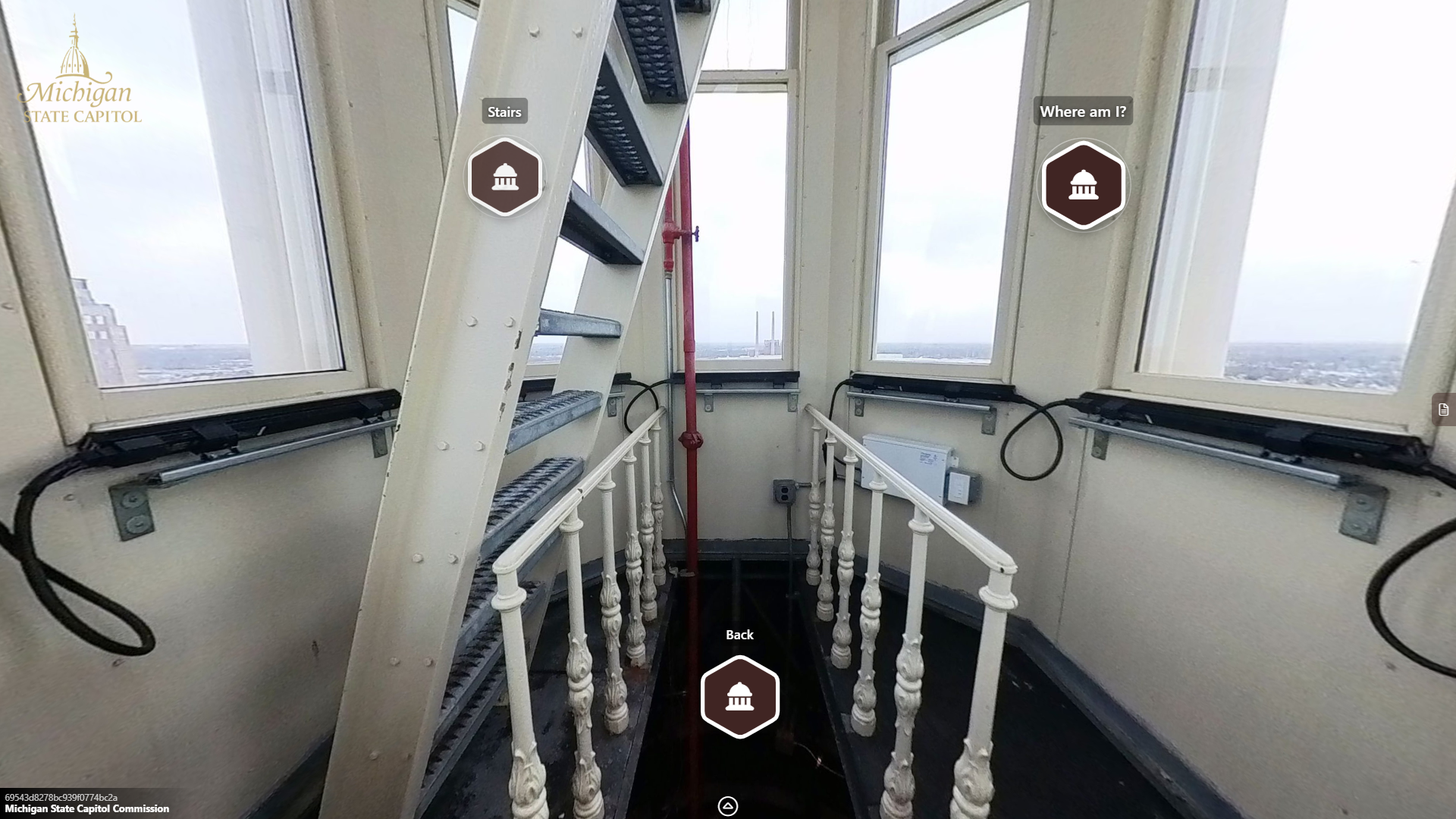 a narrow room with angled walls and tall thin windows looking outward to a city skyline. A ladder leads upward inside the room.