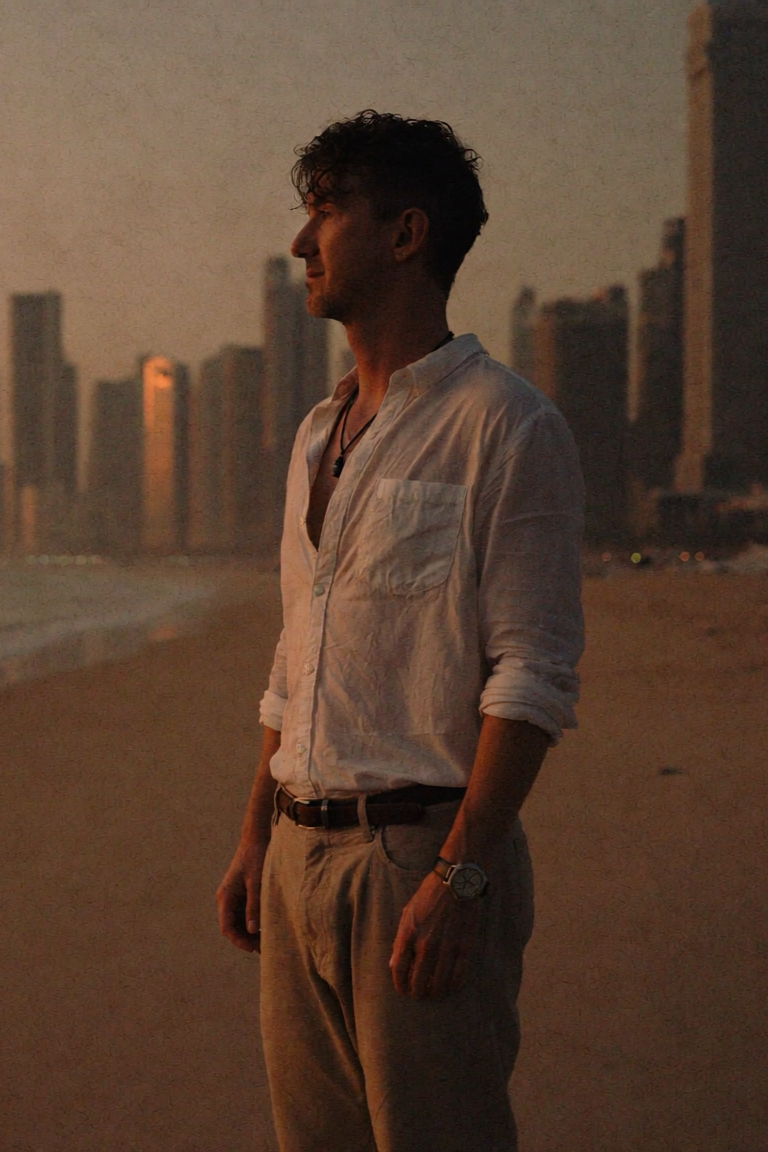 Smiling person with curly hair and tattoos, wearing a white t-shirt with a wolf design, standing on a balcony by the sea.