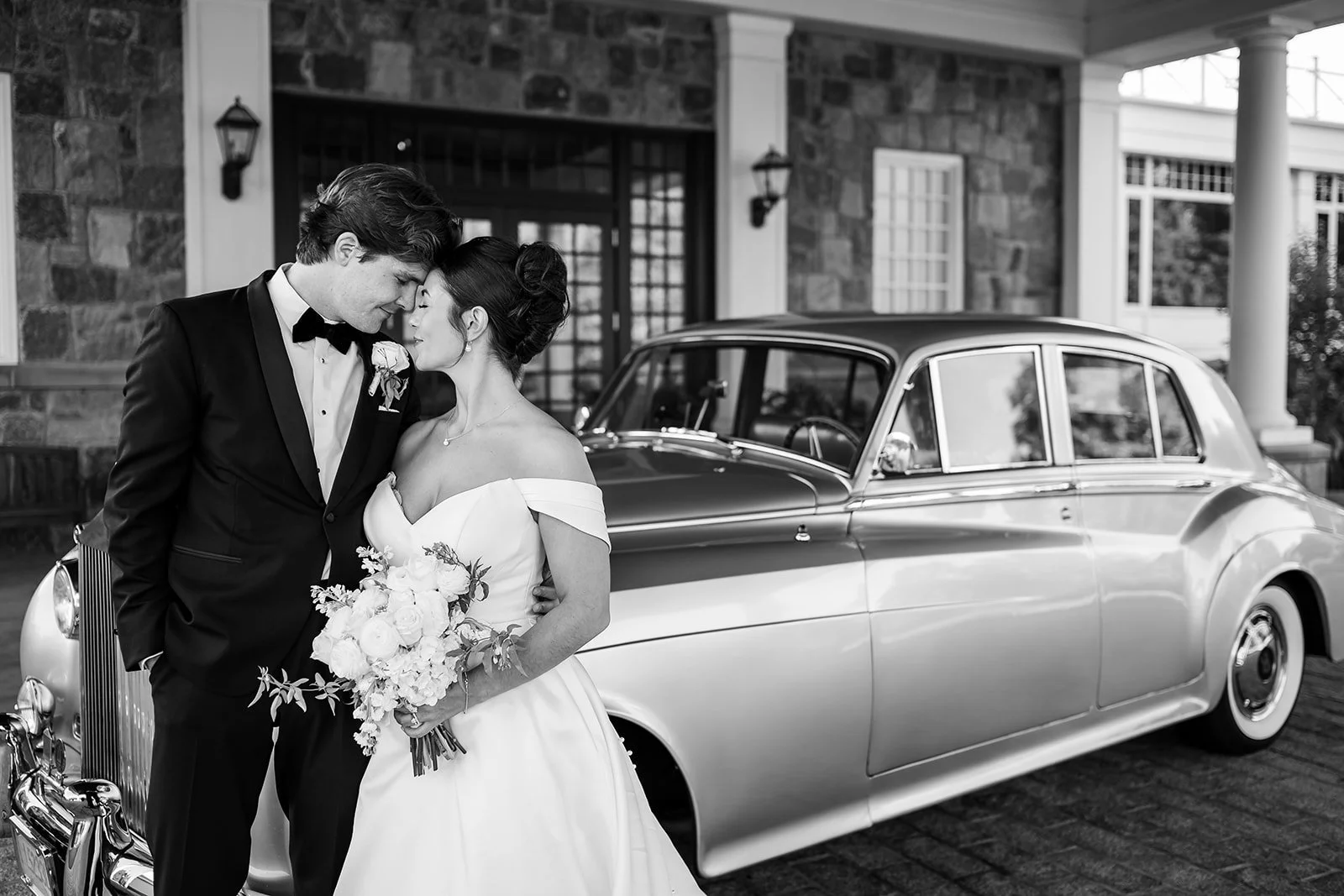 a man and woman snuggling in a tux and white gown in front of a Rolls Royce in Black and White