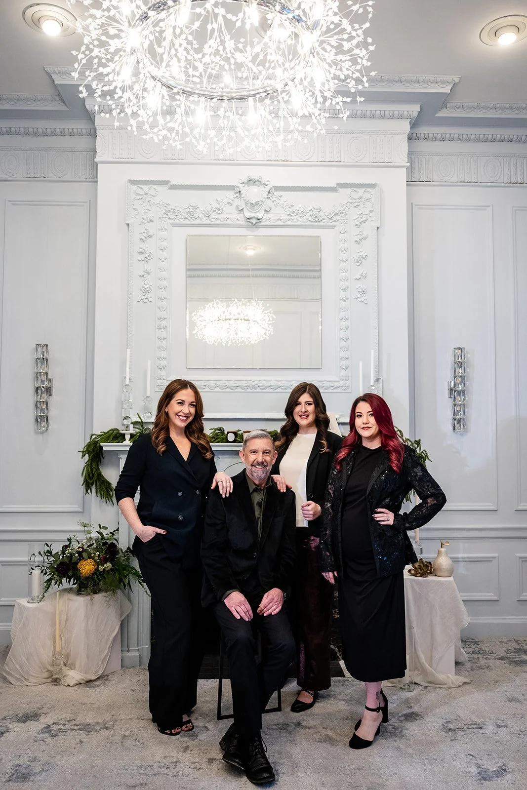 Three women in black standing around a man sitting in black by a fireplace