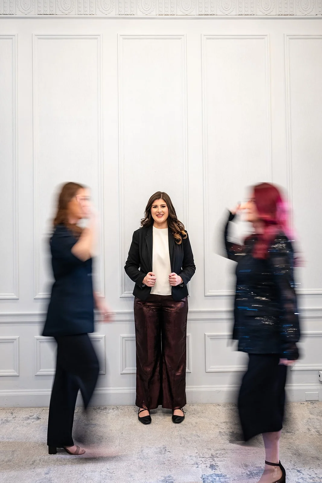 Woman standing against white wall with two blurry women walking past 